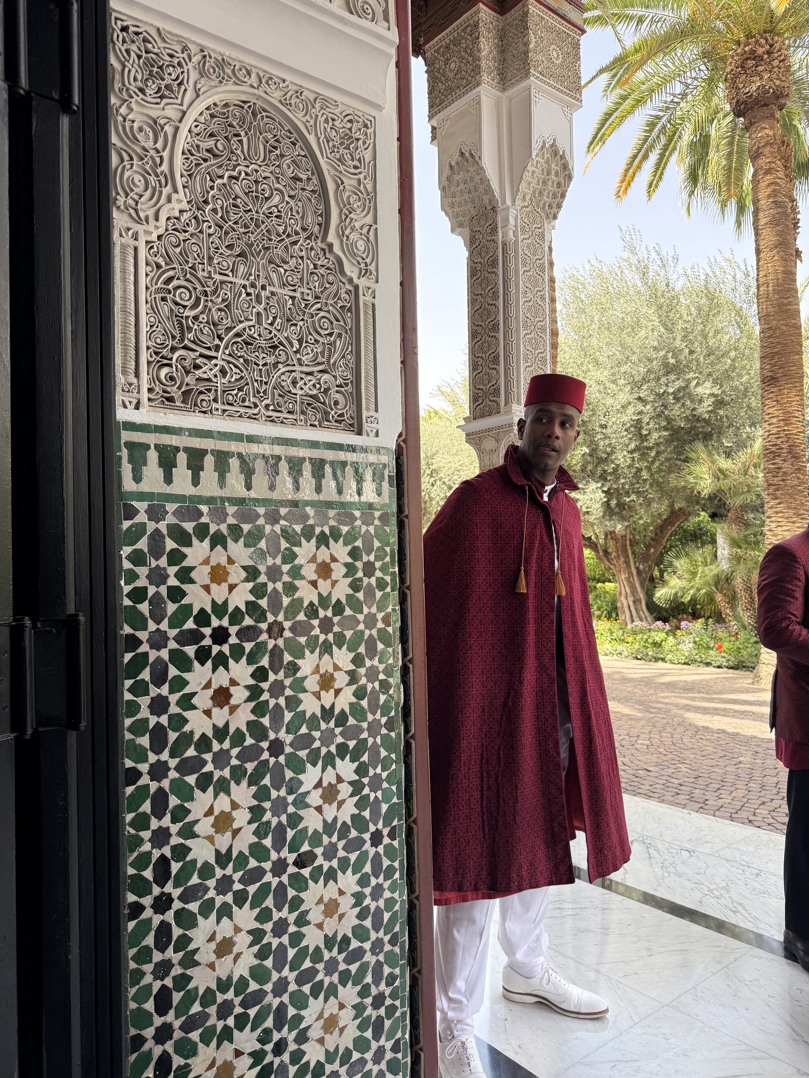 A doorman in a dark red cape and white trousers standing in an arched doorway beside zellige tiles