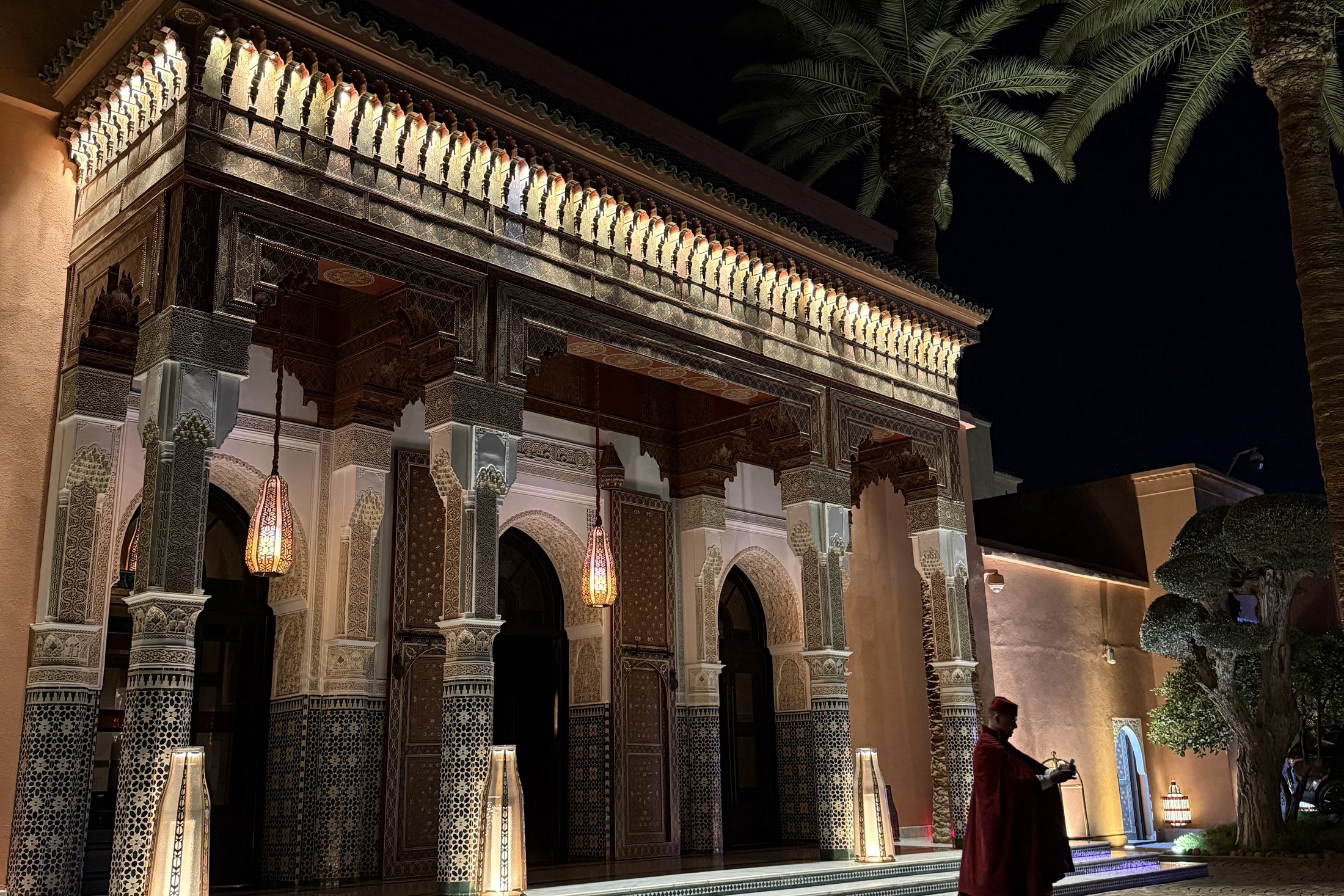 The lit entrance of La Mamounia at night, the arcades glowing and a man in a red cape crossing the forecourt
