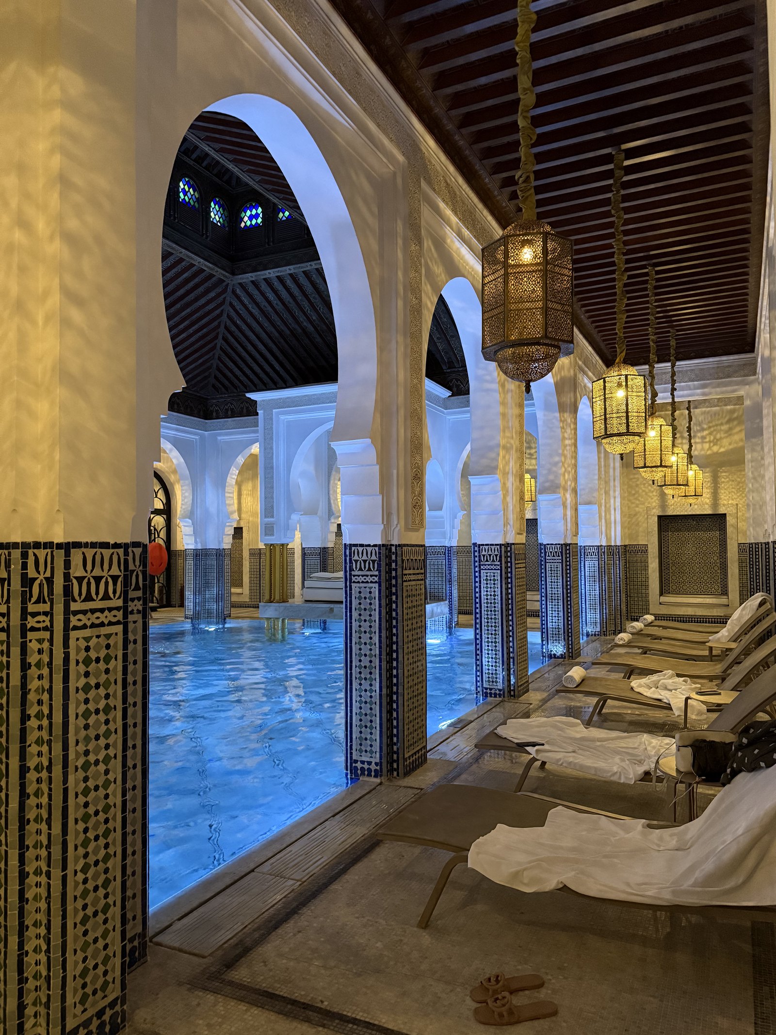 A tiled pool with a lit ceiling of arches, lanterns hanging over the edge and loungers along one side