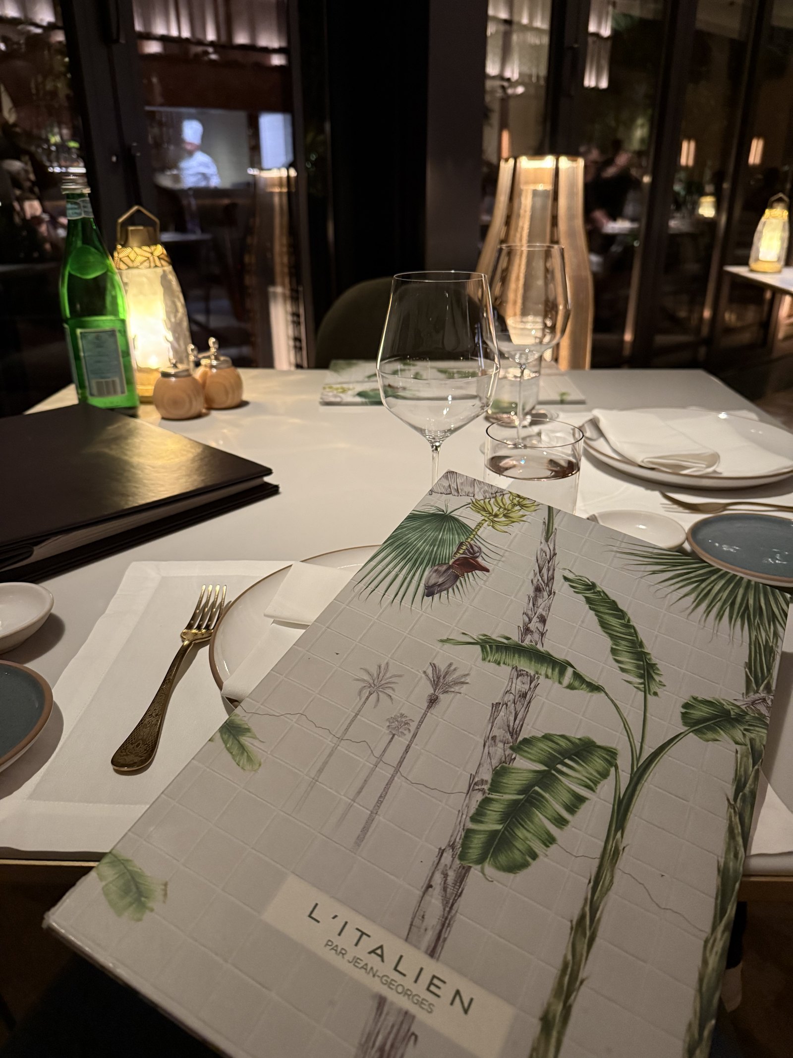 A menu printed with palm fronds on a white tablecloth, beside a glass and a folded napkin