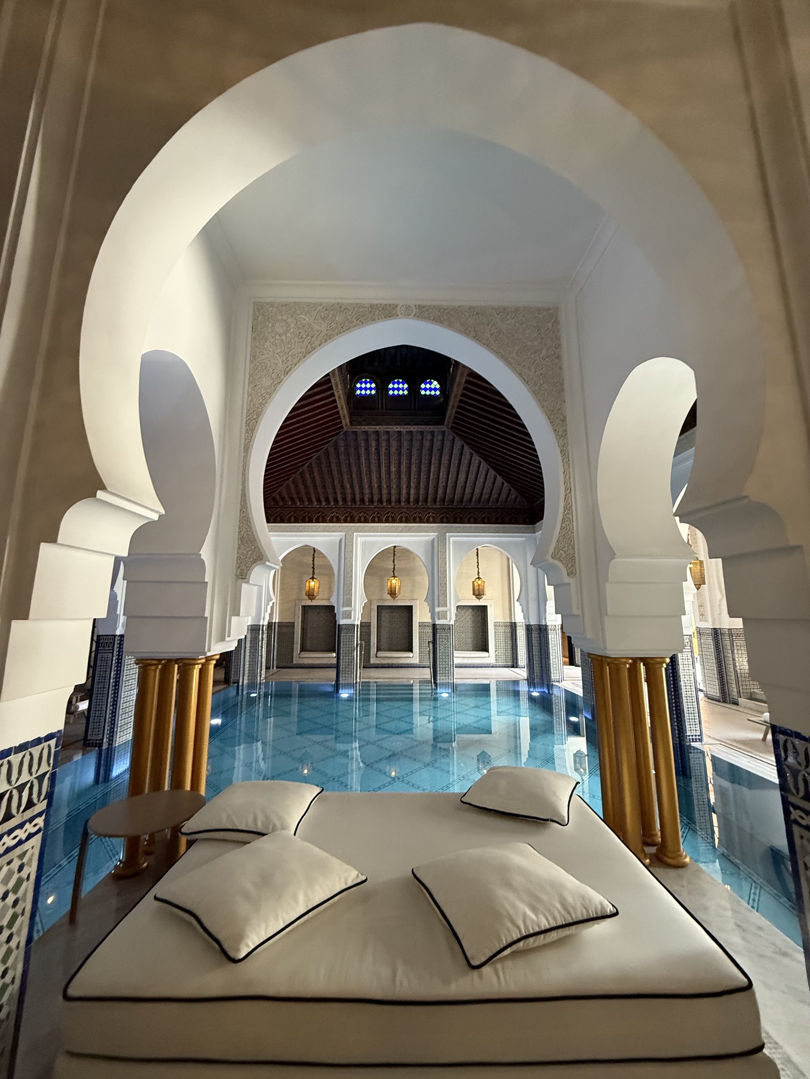 A white daybed with cream cushions set at the edge of a blue-tiled pool beneath an arch