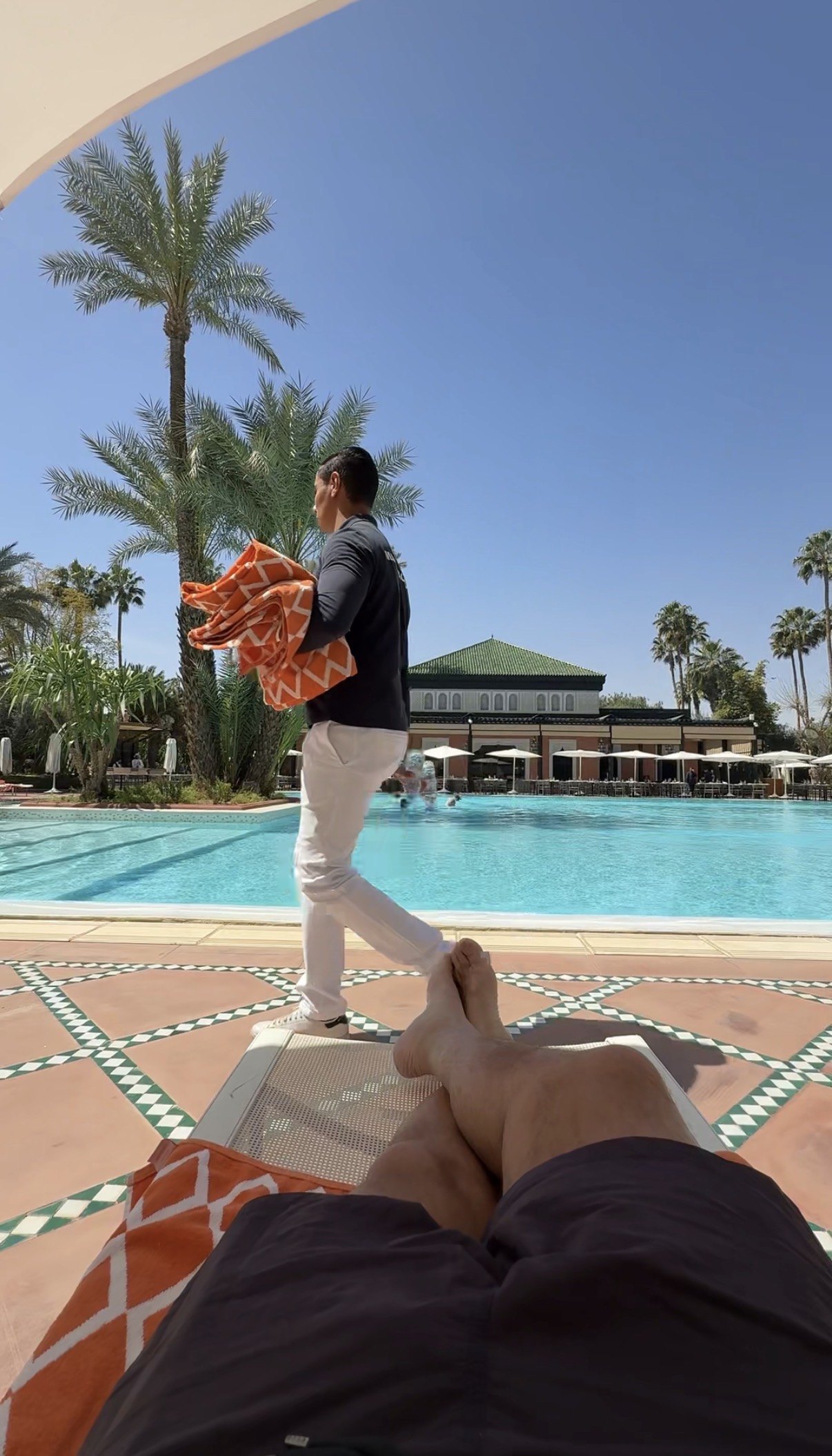 Looking down at the poolside, with a woven orange cloth on a sun lounger and bare feet resting on a pillow