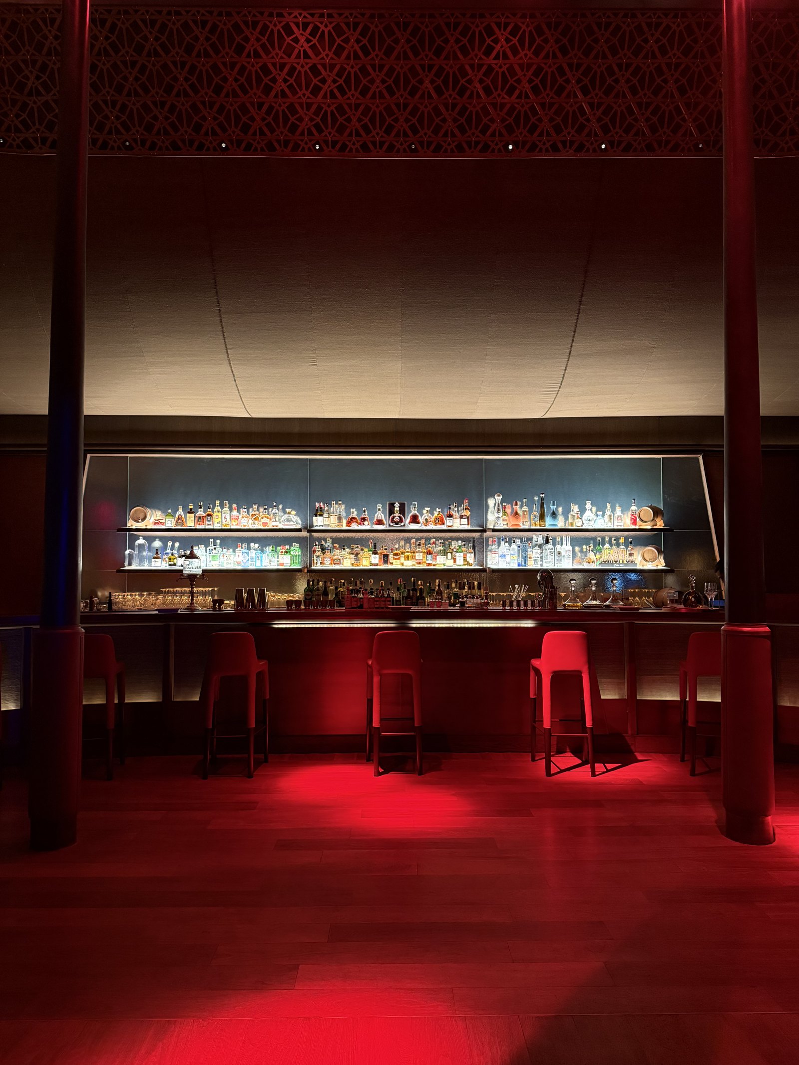 A red-lit bar with stools, a long shelf of bottles behind and a glowing counter