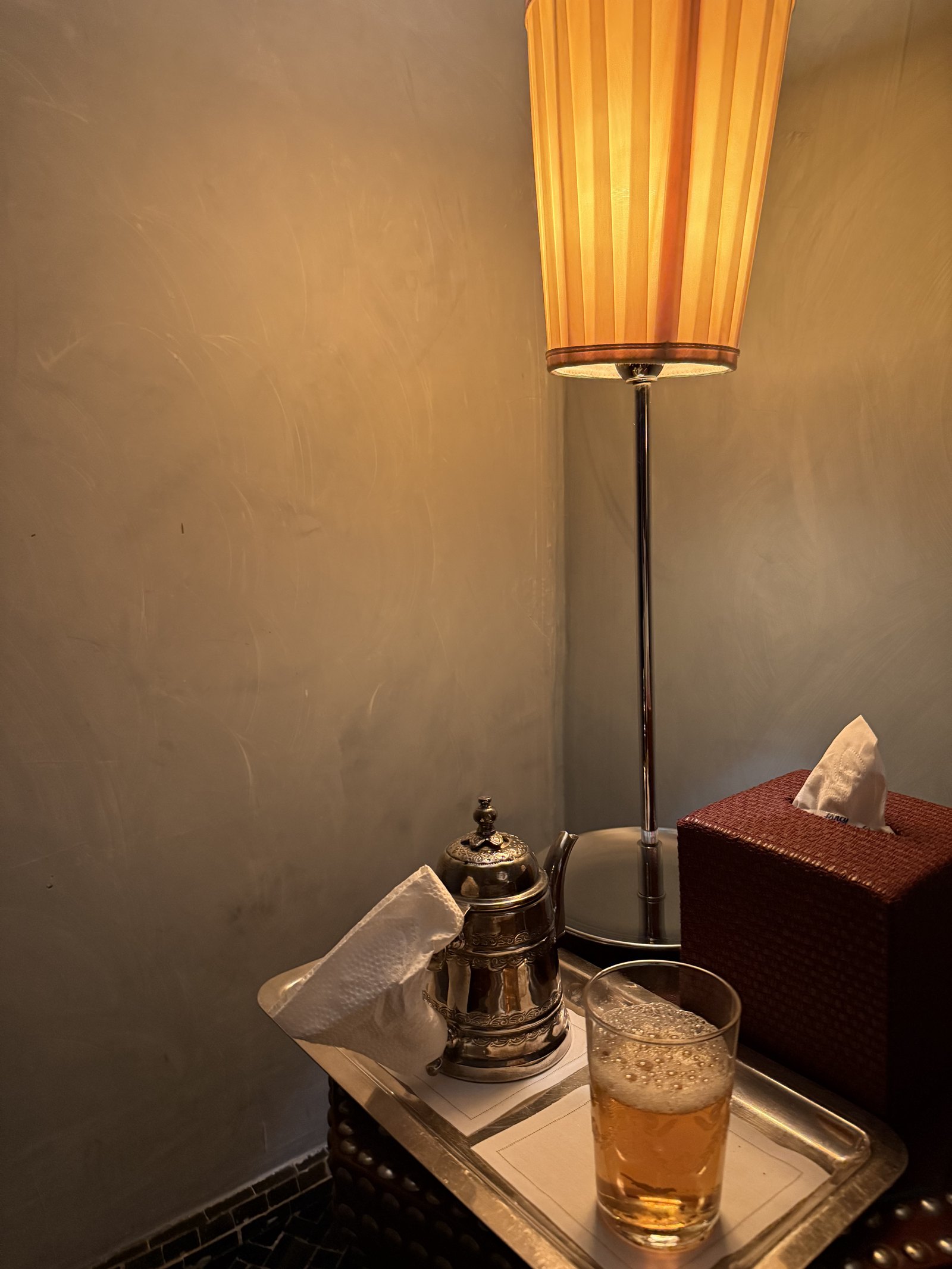 A silver teapot on a tray beside a pleated lamp and a glass of mint tea