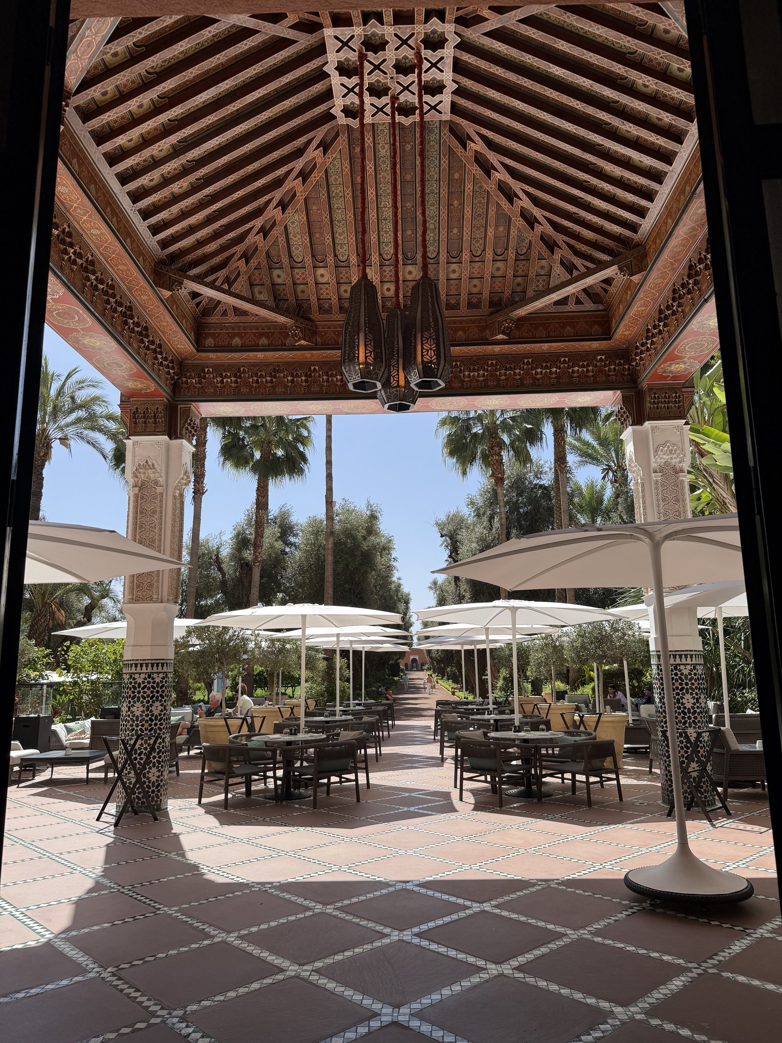 A wooden pavilion with a carved ceiling and white parasols over a terrace of tiles