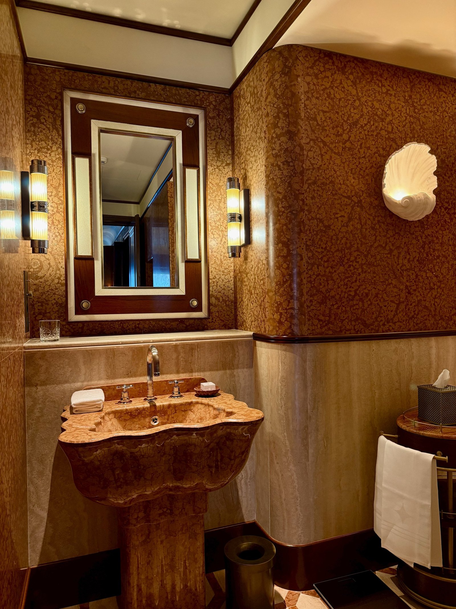 A burl wood basin under a deco mirror with a shell sconce beside it