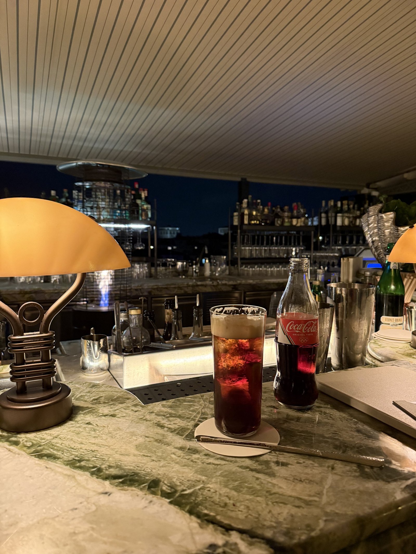 A tall dark drink and a Coca-Cola bottle on the rooftop bar at night