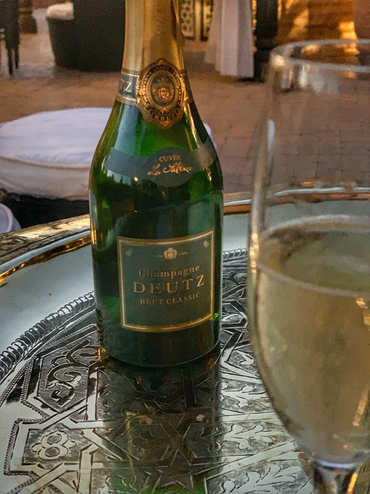 A bottle of Deutz Brut Classic with a Cuvee La Sultana neck collar and a filled glass on a chased silver tray
