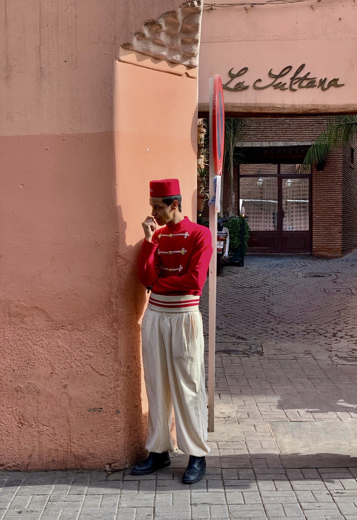 A doorman in a red jacket and white trousers standing against the pink wall beside the La Sultana entrance in the Kasbah
