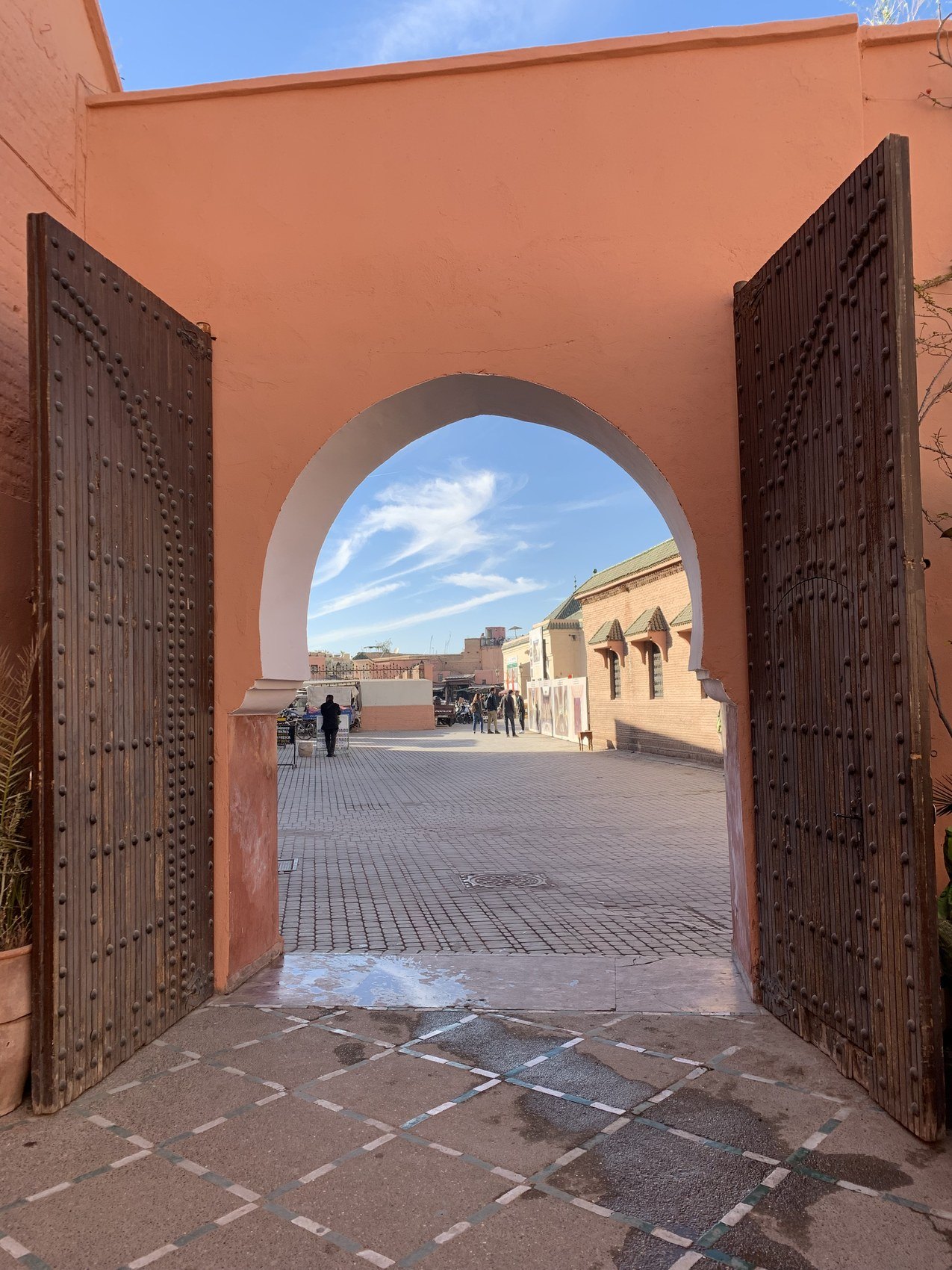 A pair of studded timber doors standing open under a horseshoe arch onto a sunlit square in the Kasbah