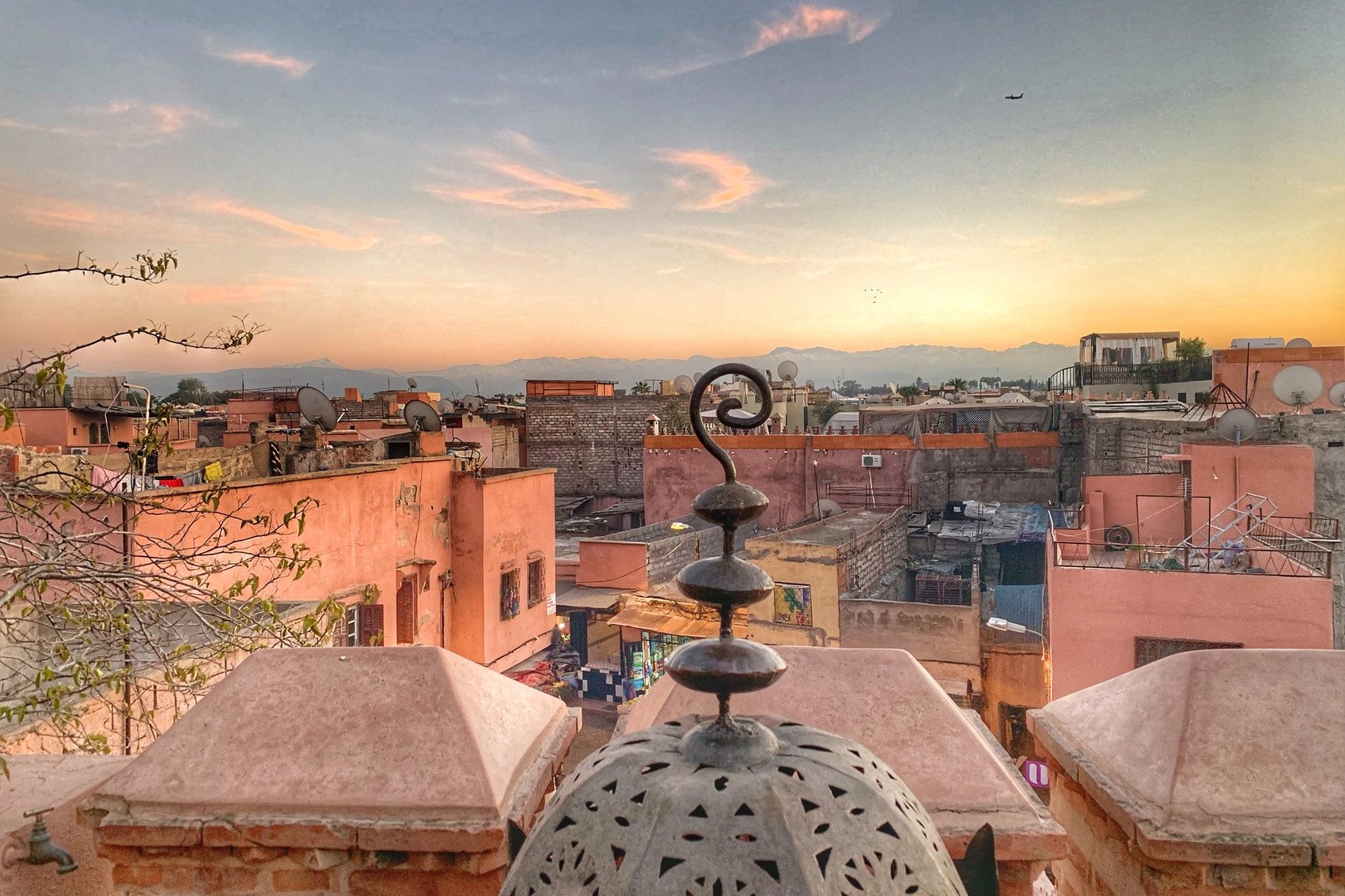 The rooftops of Marrakech at sunset seen from the terrace at La Sultana, with a pierced lantern finial in the foreground and the Atlas in the distance