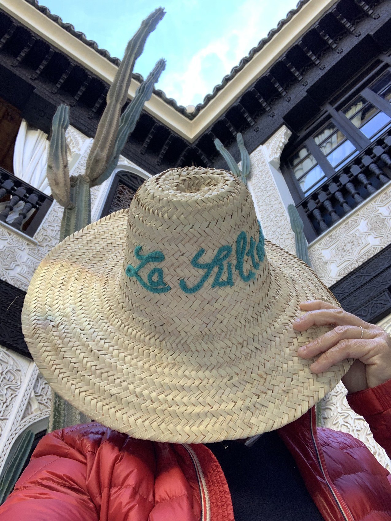 A wide straw hat embroidered La Sultana in green thread, held up against a courtyard and a blue sky