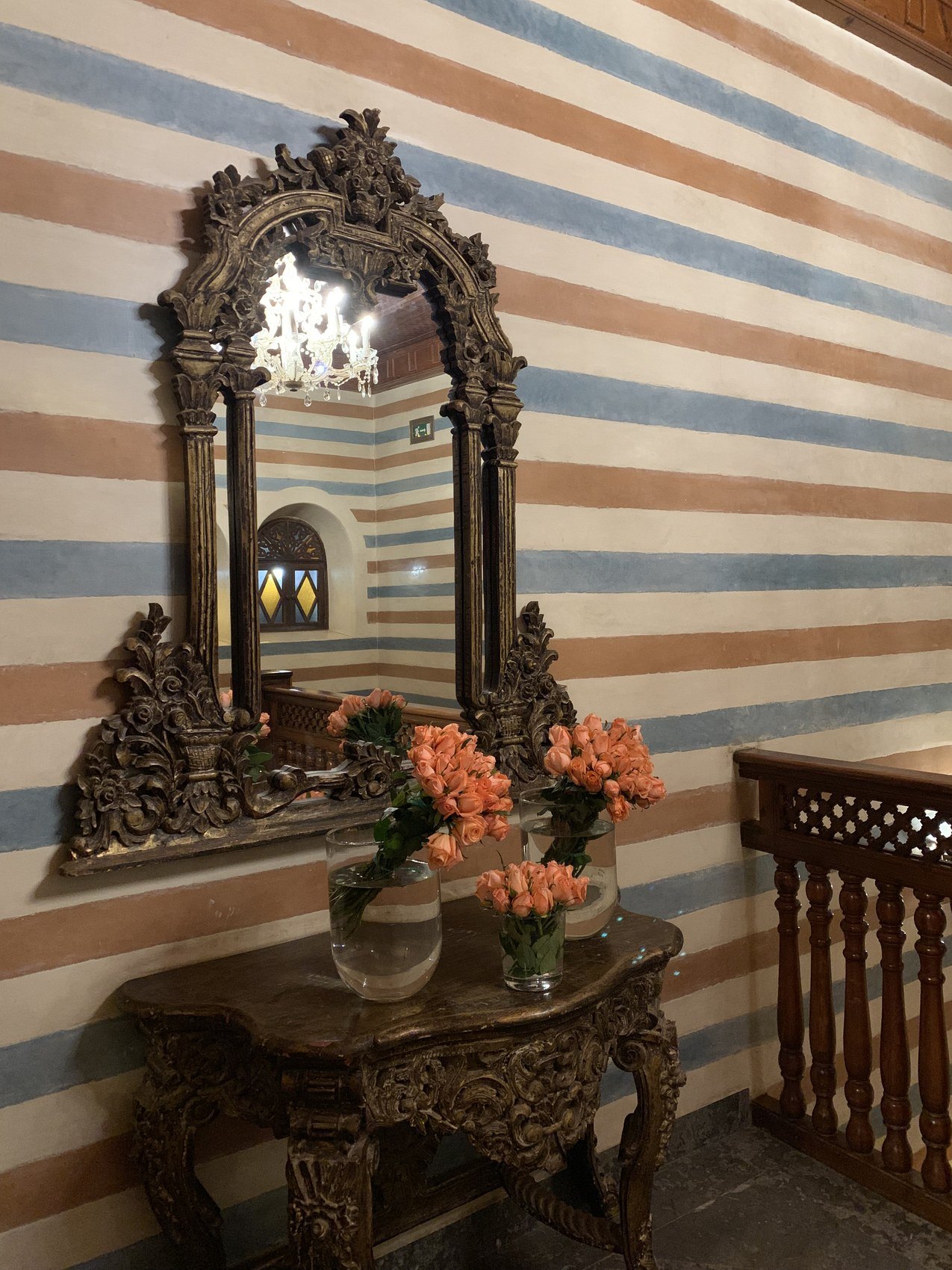 A carved gilt mirror and a console with coral coloured flowers against a blue and white striped wall at La Sultana