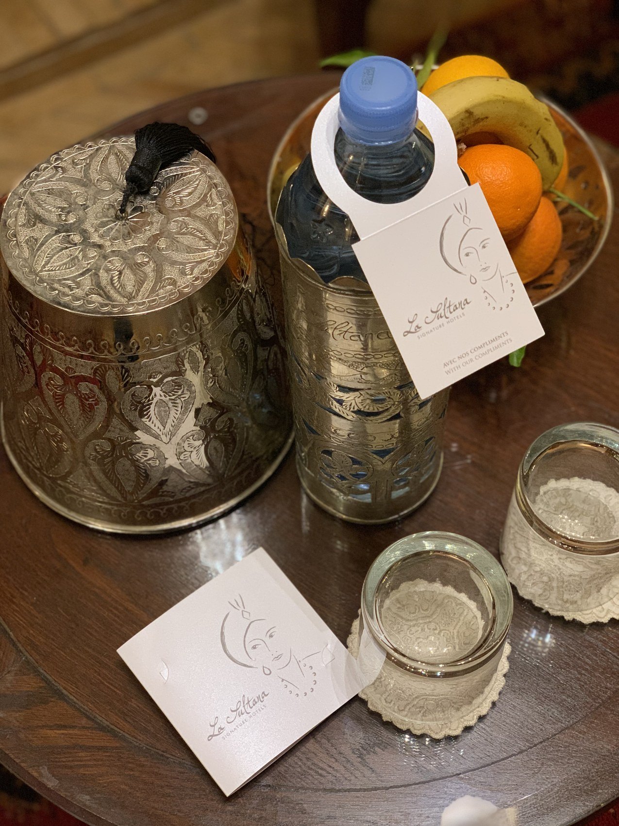 A chased silver box, a water bottle with a La Sultana tag and two handwritten cards on a dark table