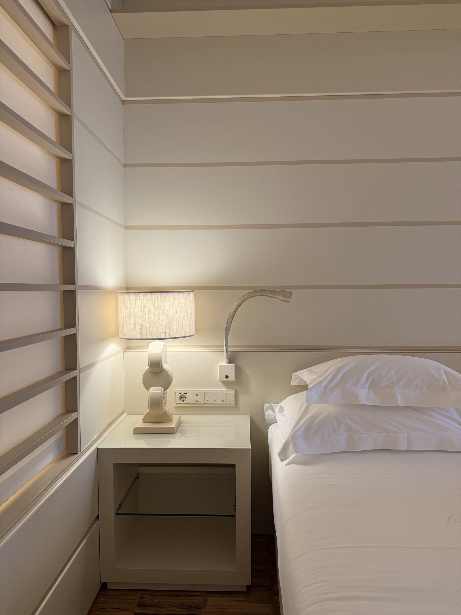 A bedside table with a lamp and a reading light, beside white linen and a slatted wall