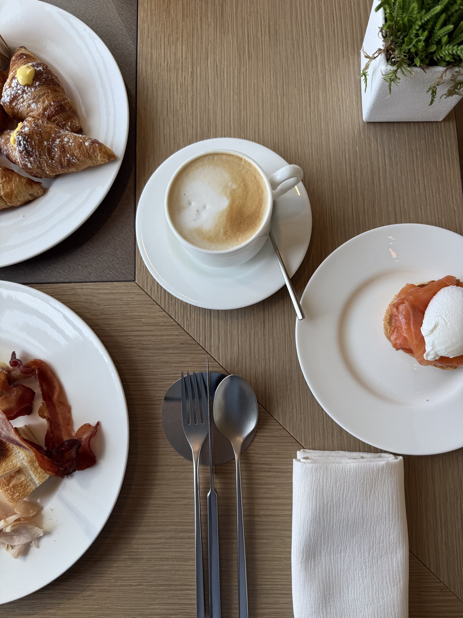 A breakfast table from above with croissants, a cappuccino, bacon and poached egg on salmon