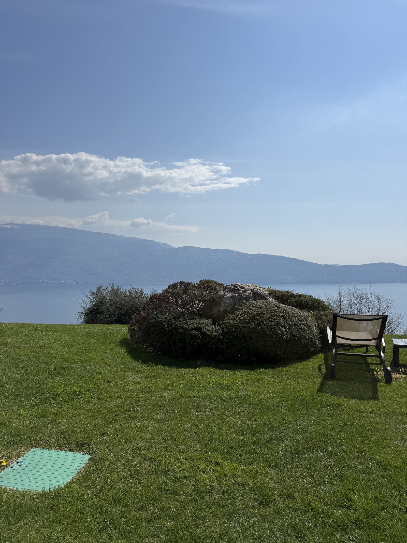 A single chair on a lawn beside a clipped bush, with the lake and the mountains beyond