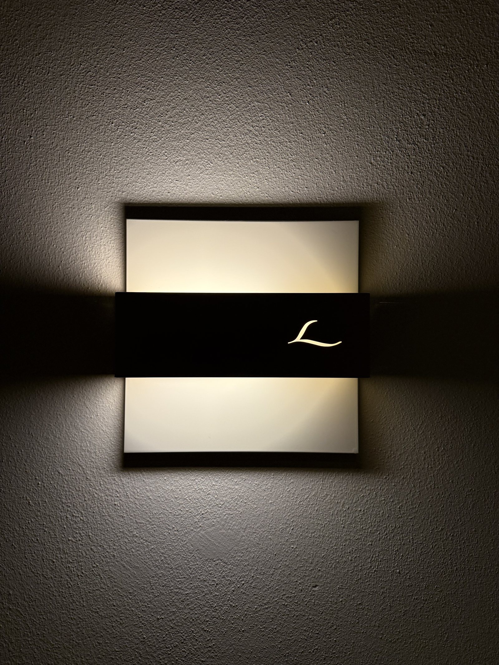 A wall light in black and white with the Lefay L cut into it, lit from within