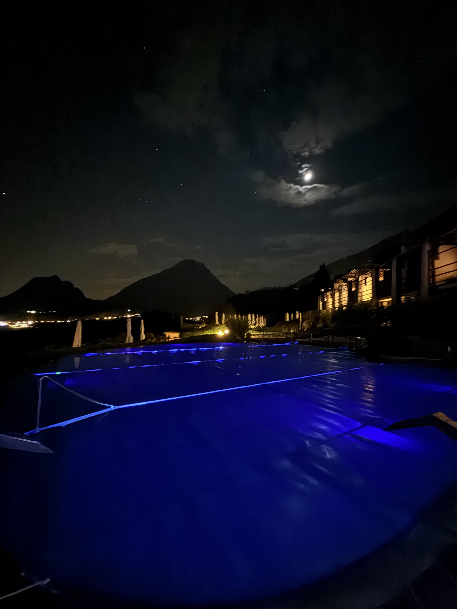 The outdoor pool lit blue at night under a clouded moon, with mountains beyond