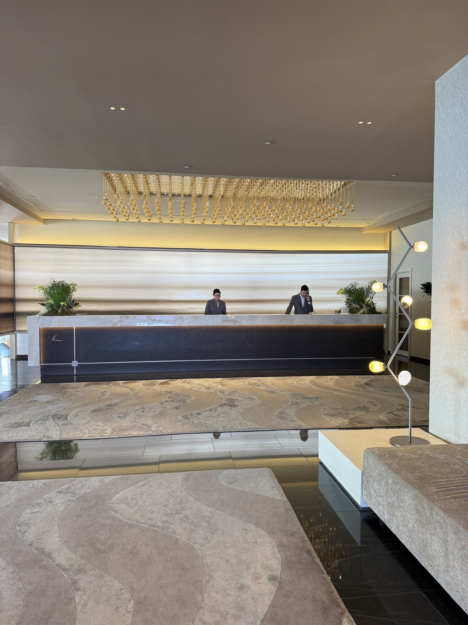 A long dark reception desk under a rectangular chandelier, on a floor of veined stone