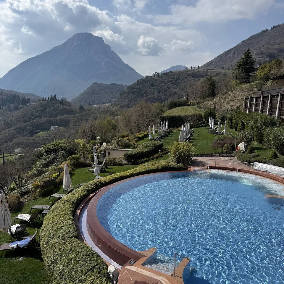 A circular outdoor pool set into terraced lawns above the lake, with olive trees around it