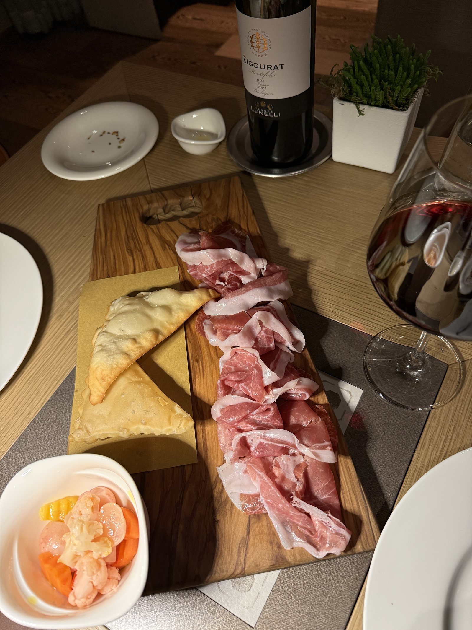 A wooden board with cured meats and flatbread, a bowl of prawns and a bottle of red wine