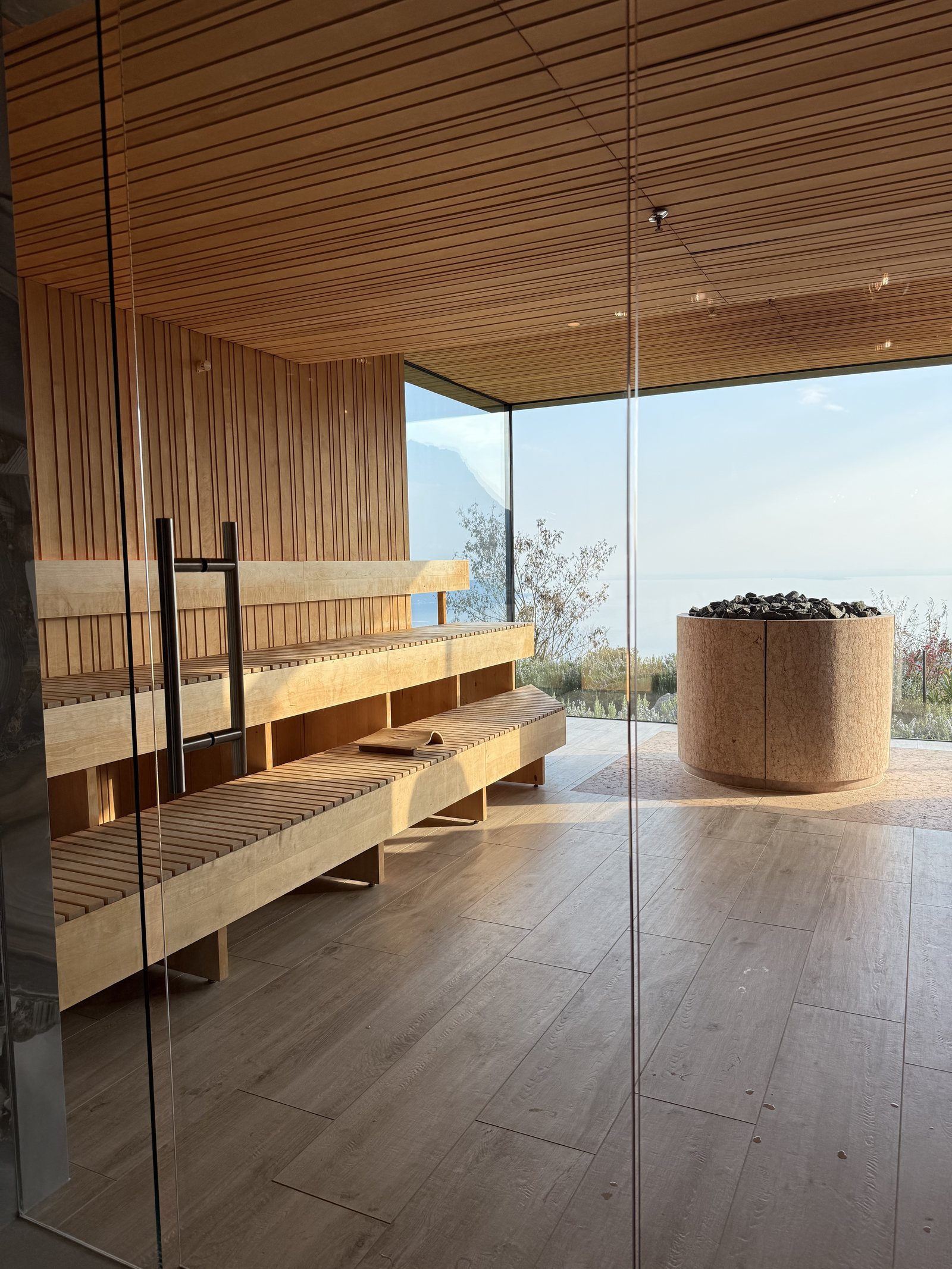 A sauna in pale timber with tiered benches and a stove of dark stones behind glass