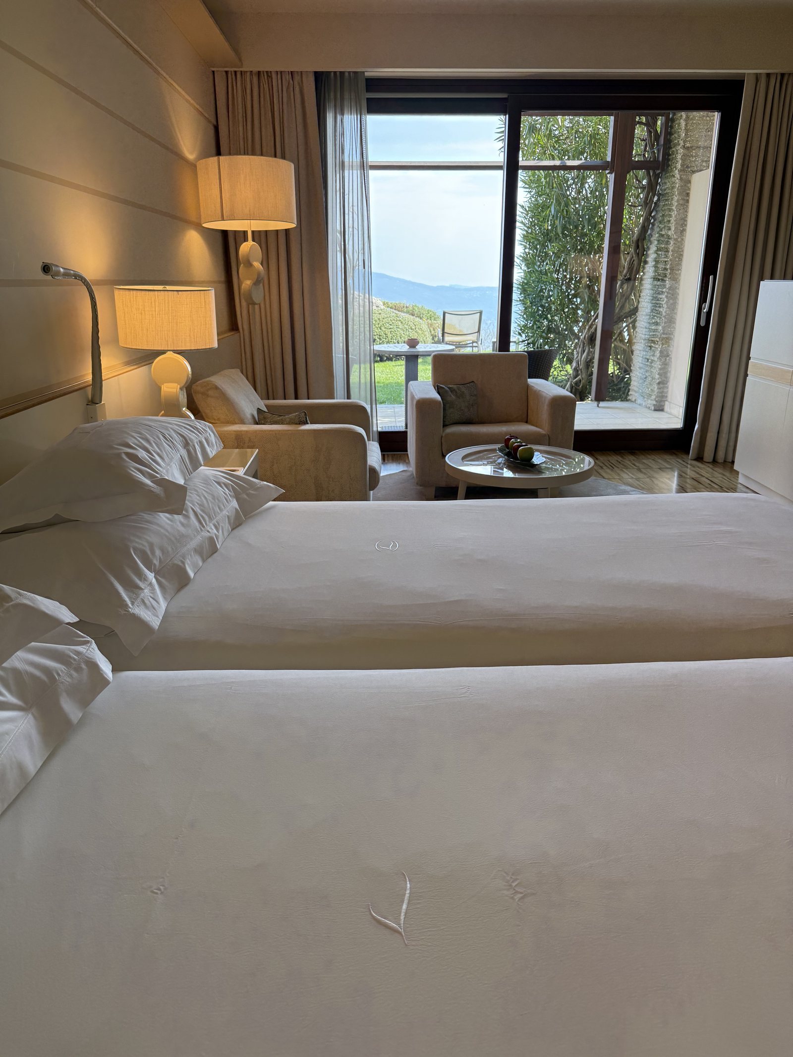 A made bed seen from the foot, with a window beside it framing trees and the lake
