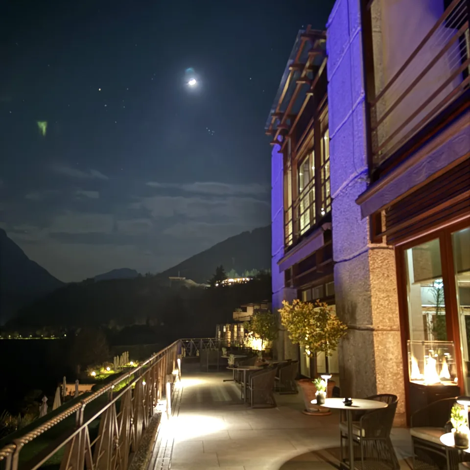 A lit terrace at night with small tables, the mountains dark behind it under a moon