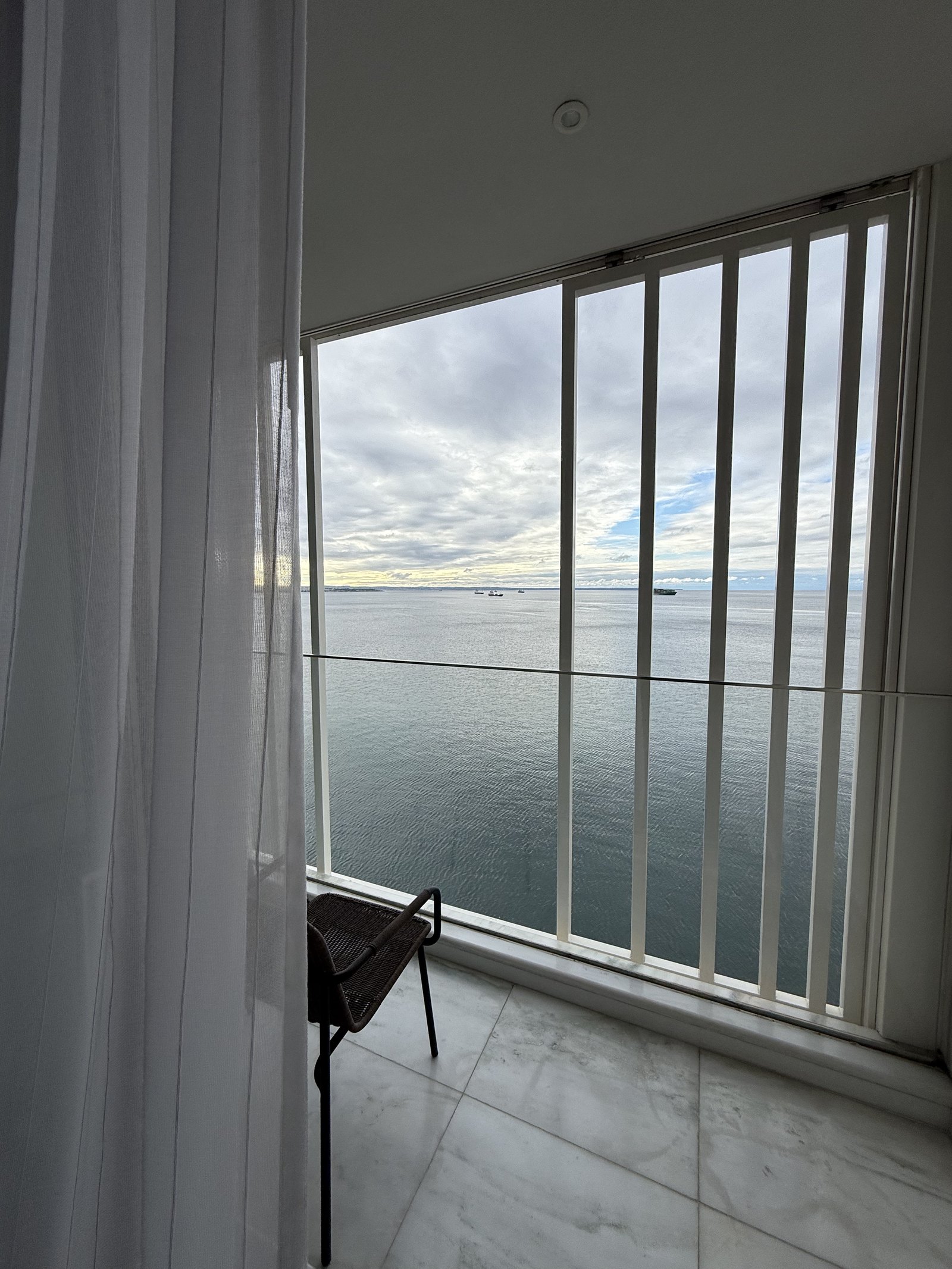 A small balcony with a chair and a table behind tall glass, the flat grey water of the gulf beyond