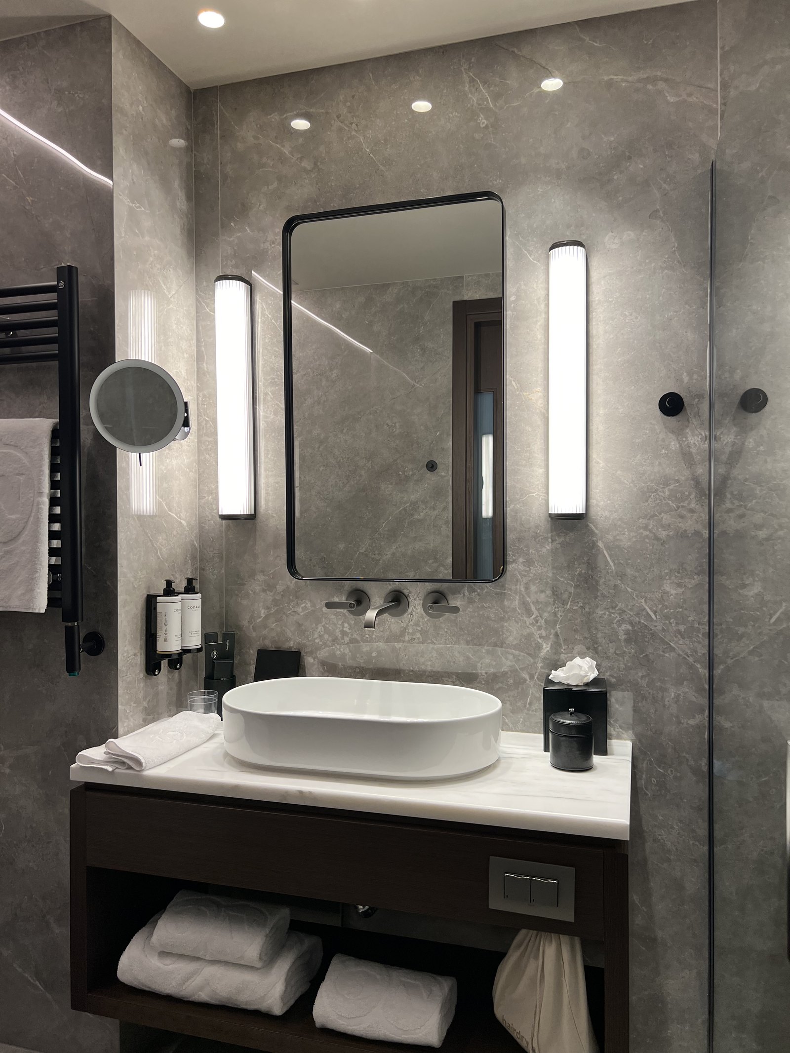 A bathroom in grey marble with an oval basin on a dark vanity, a mirror between two vertical light tubes and a heated towel rail