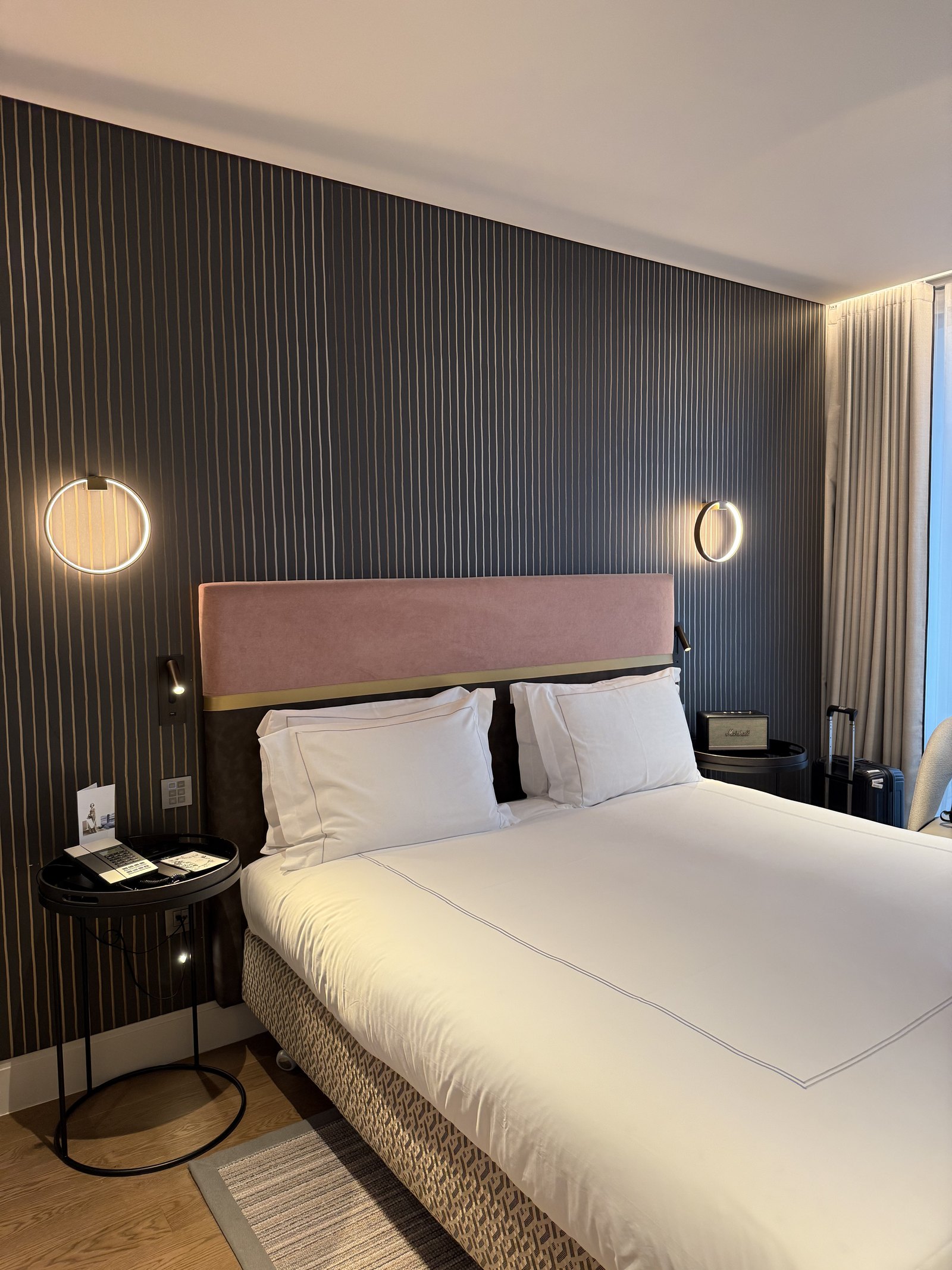 A bed with a rose-coloured headboard against fluted grey panelling, round wall lights either side