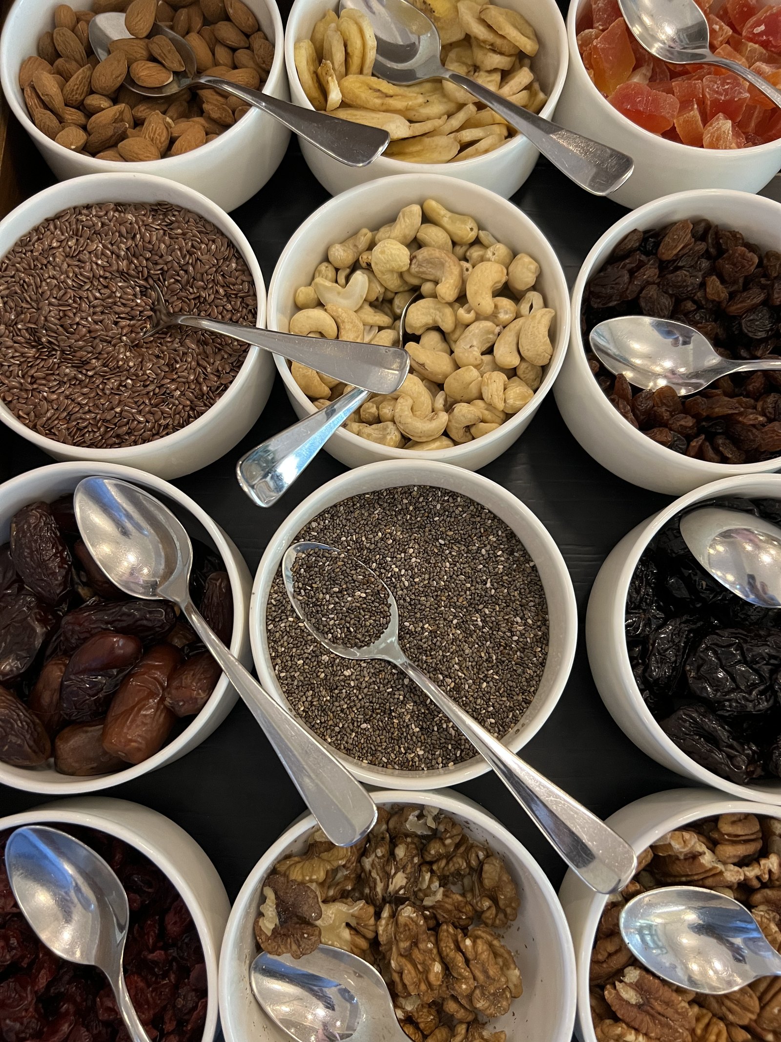 A tray of white bowls from above holding almonds, cashews, walnuts, dates, raisins, chia and dried fruit, a spoon in each