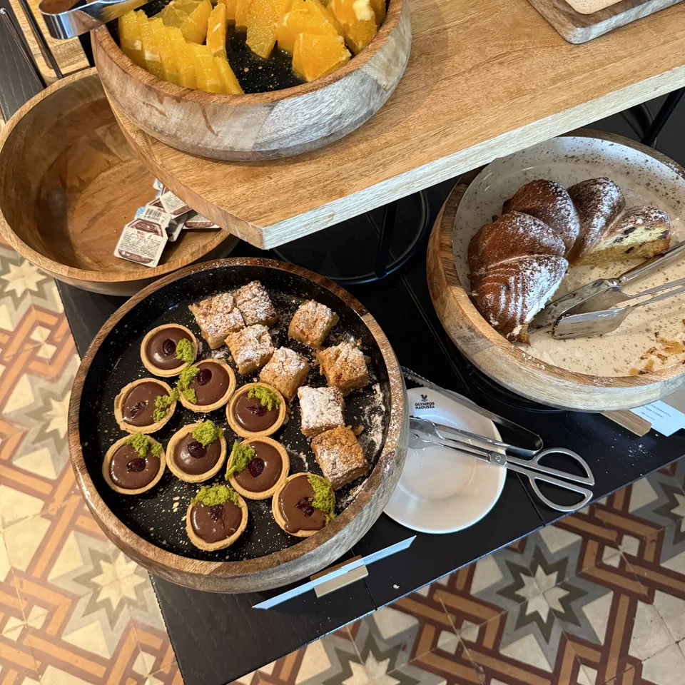 A breakfast buffet of pastries, fruit and cakes on a wooden counter above a patterned tile floor