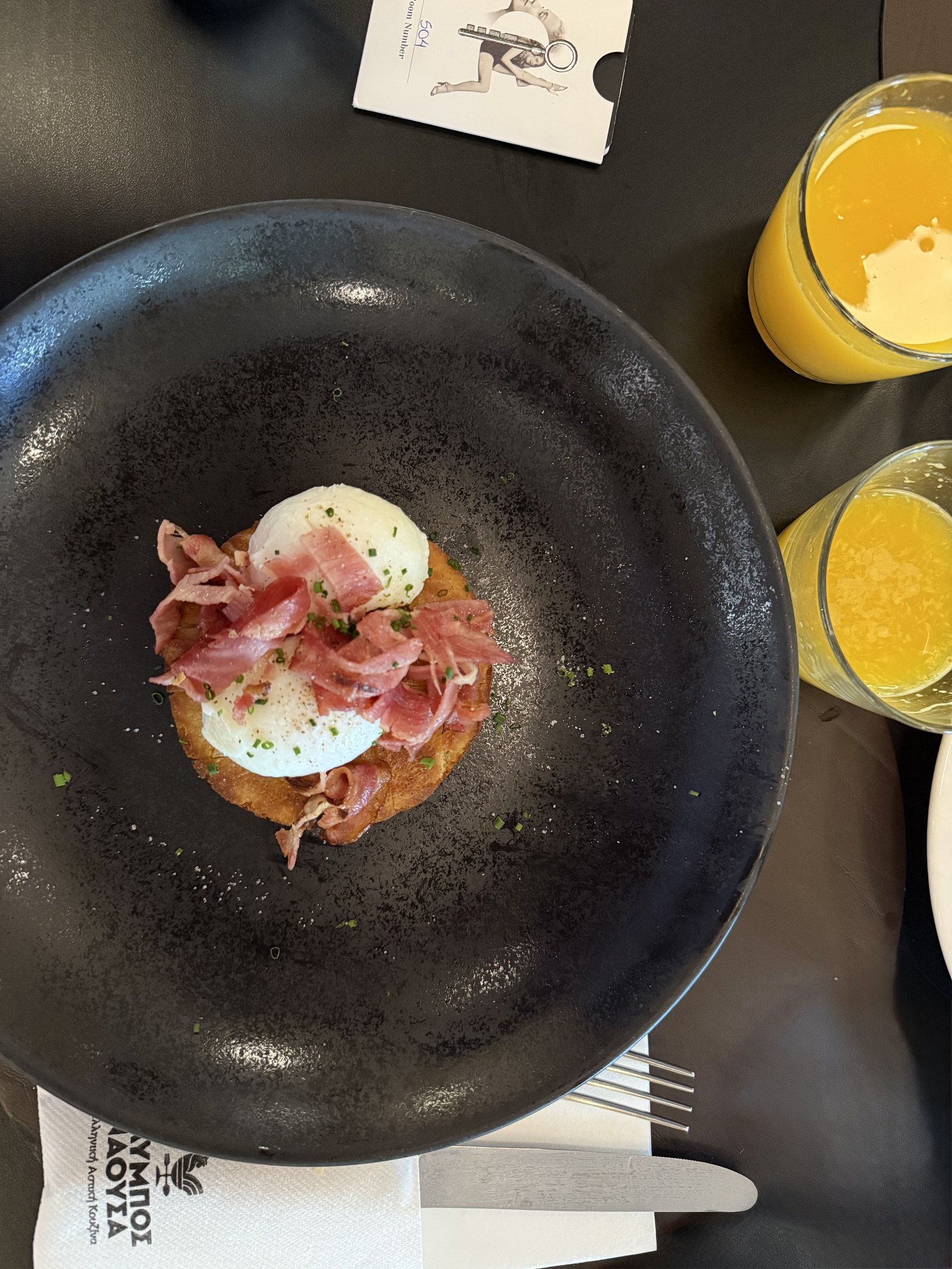 A poached egg on toasted bread with ham and herbs on a dark plate, a glass of orange juice beside it