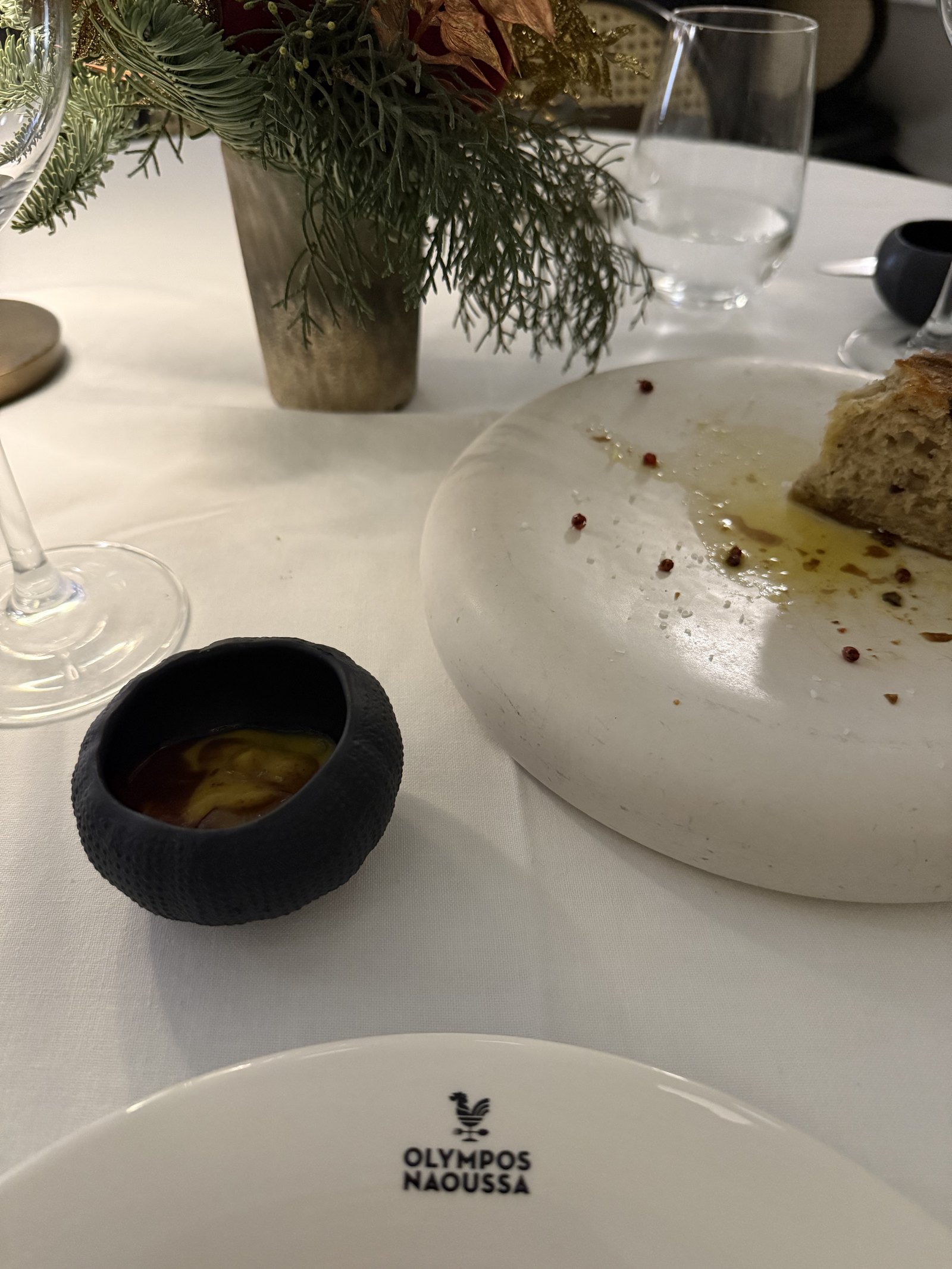A table laid in white linen with a vase of winter foliage and berries, plates and a dark bowl of sauce