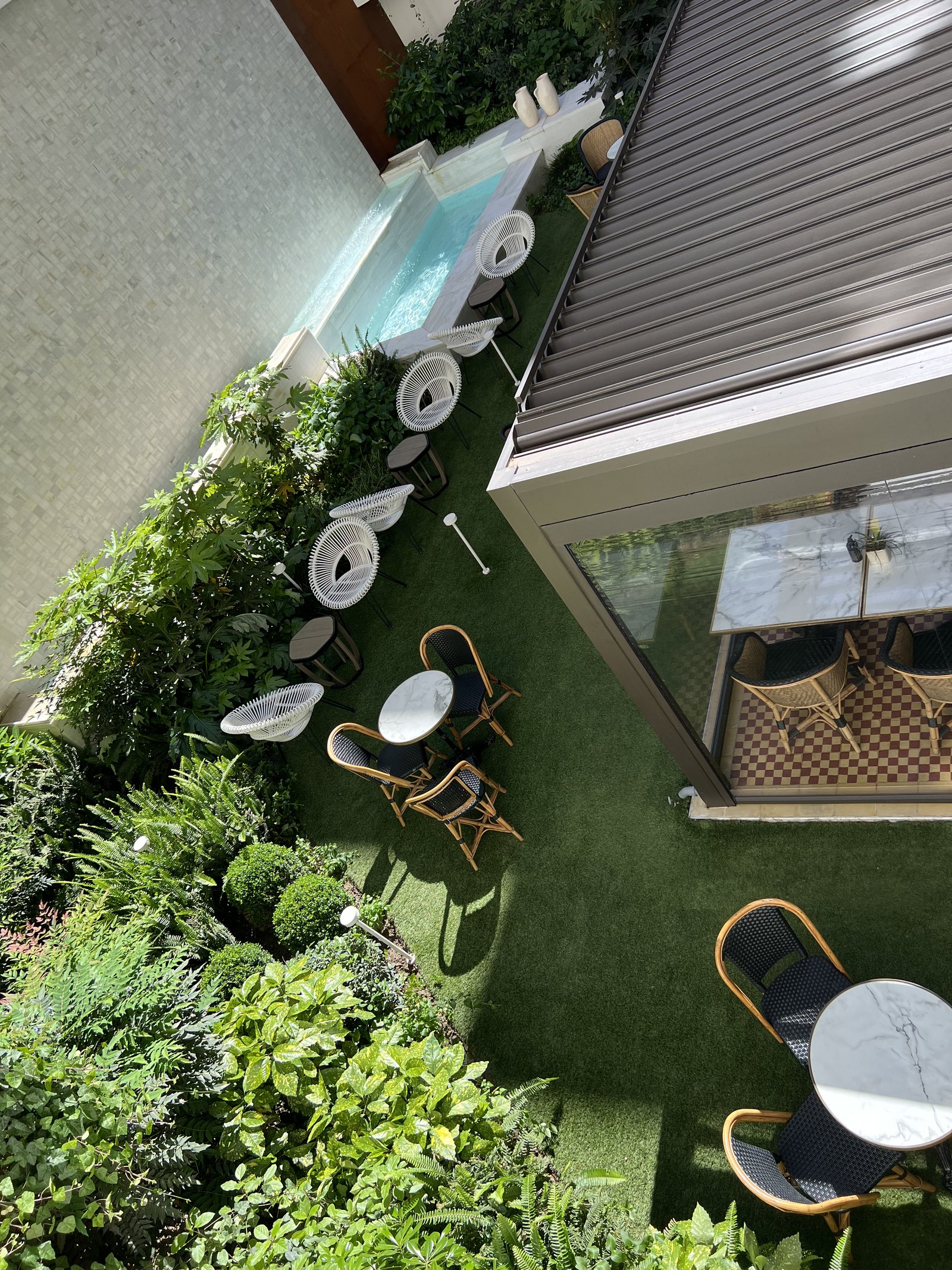 A courtyard from several floors up, a green planted wall down one side, a glass-sided water feature and cane chairs on grass