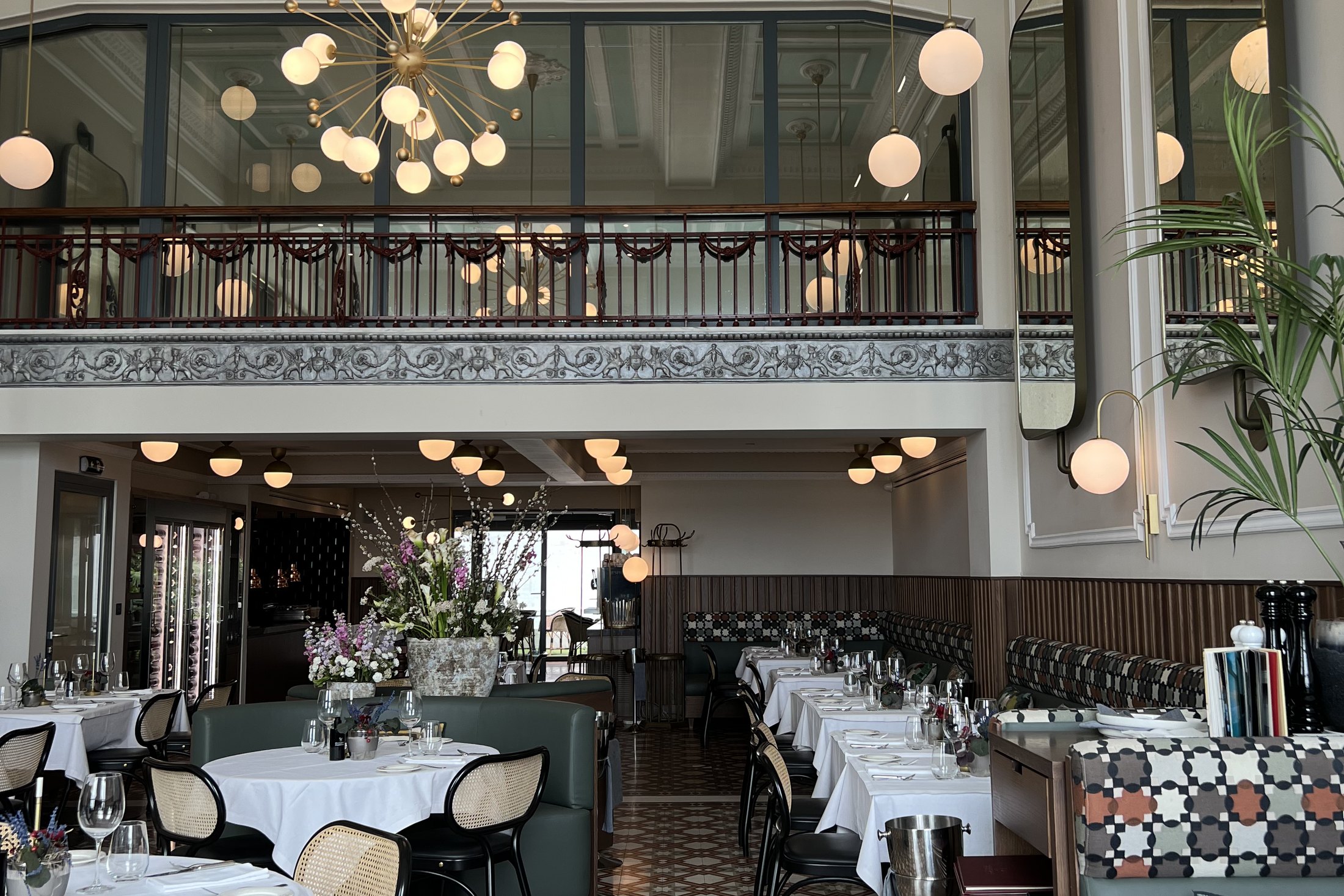 A restored 1920s dining room with a pale green coffered ceiling, a mezzanine with ironwork railings, a sputnik chandelier and glass globes over laid tables