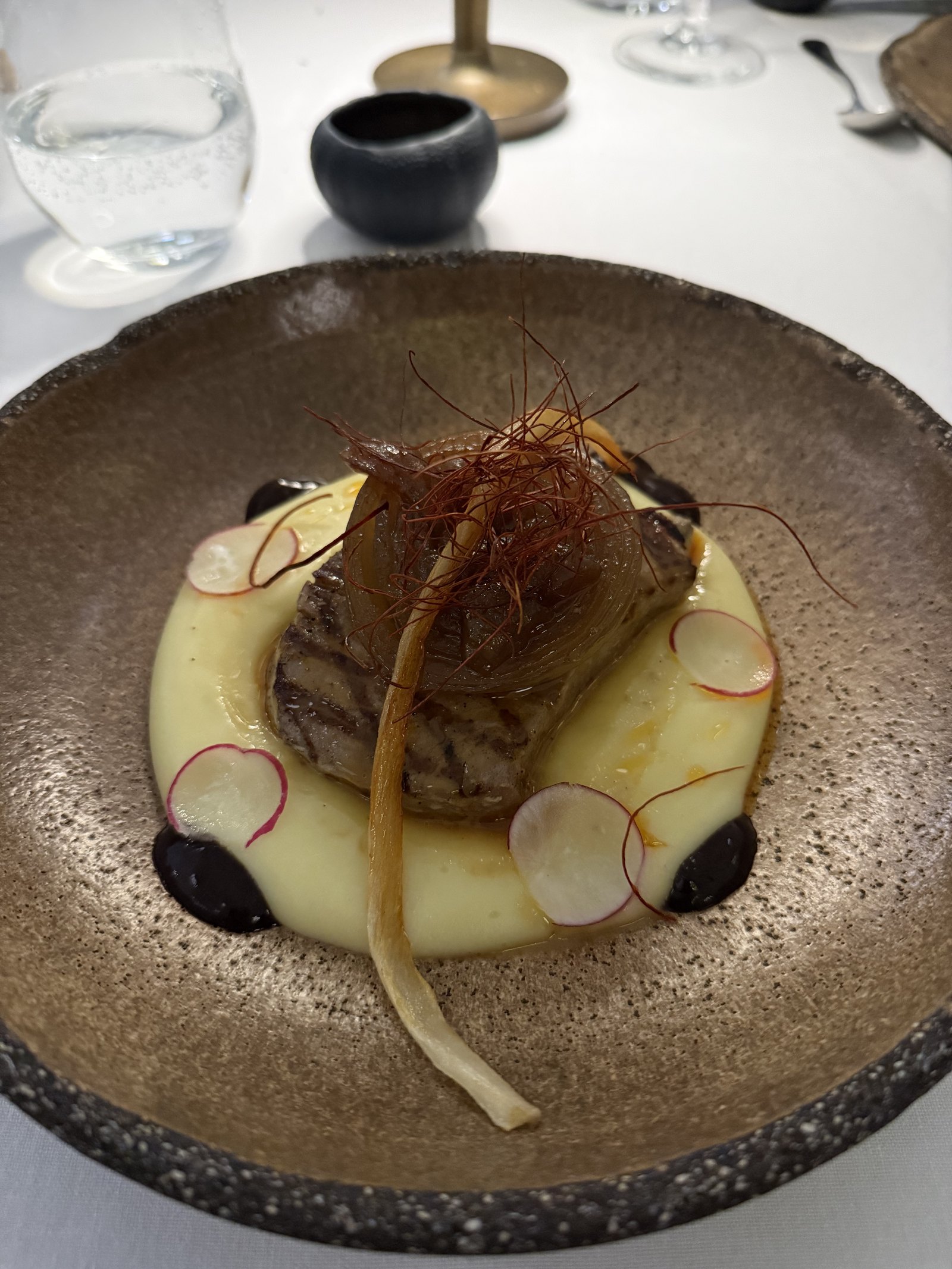 A dish of braised meat on a pale puree with radish slices and a crisp garnish on a speckled plate