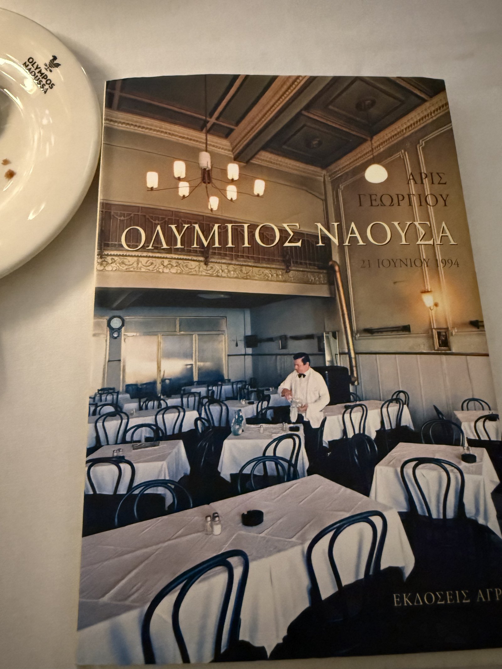 A hardback book about the history of Olympos Naoussa lying on a laid table, its cover showing the old dining room