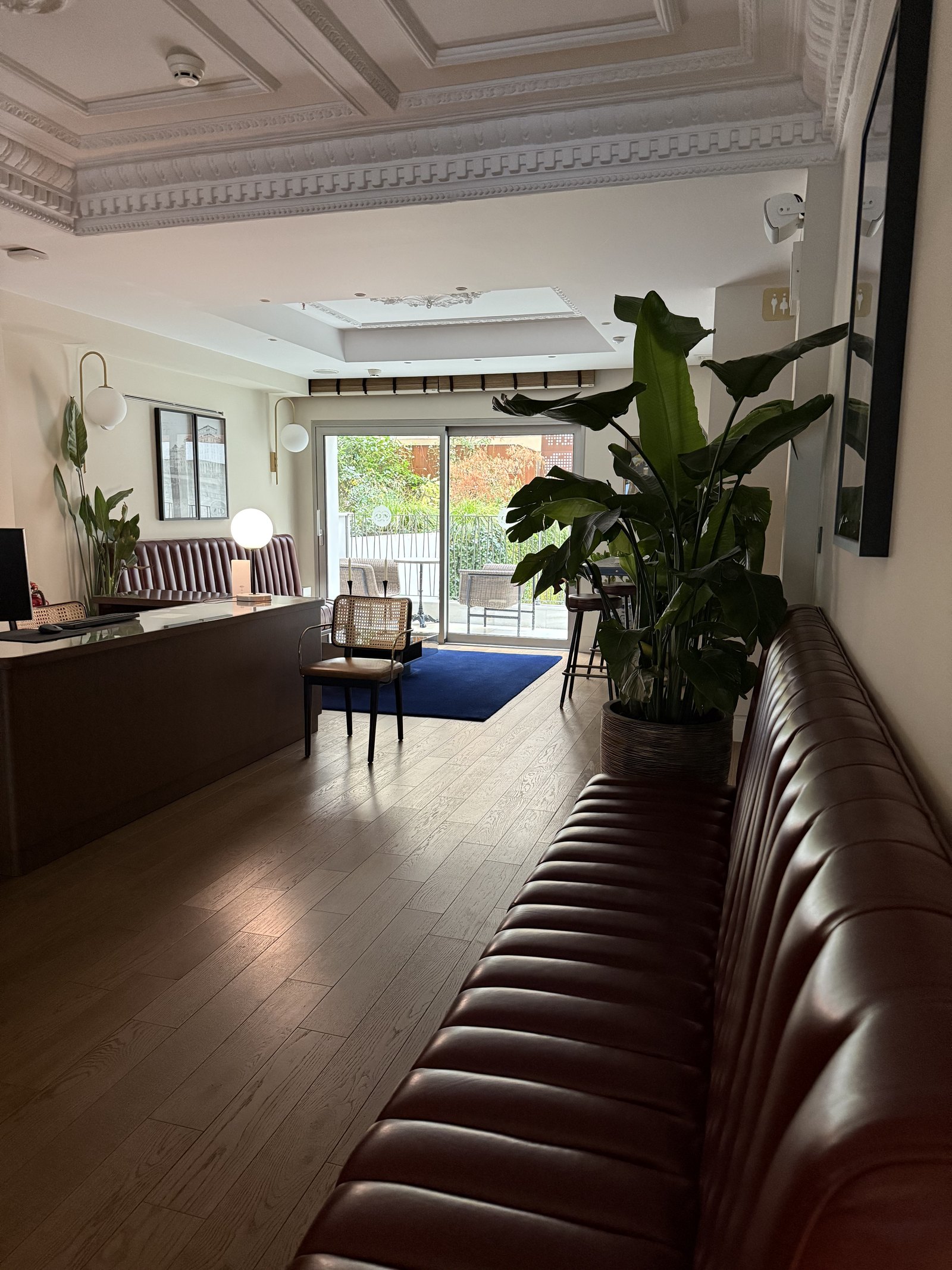 A lobby with a long oxblood leather banquette down one side, a reception desk at the far end and a tall plant beside a glazed door onto a courtyard
