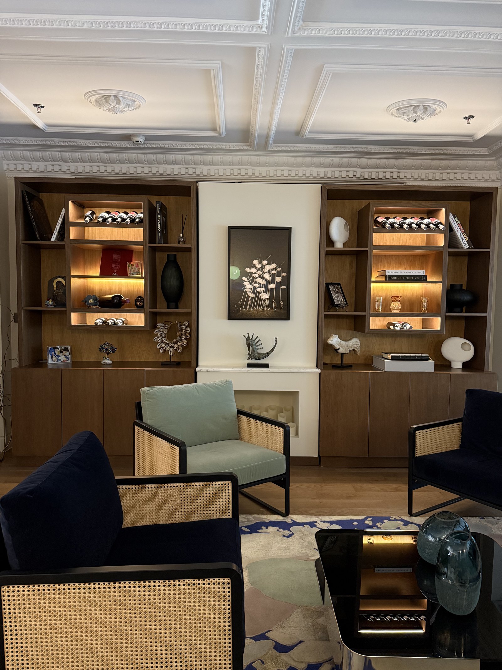 A sitting room with lit timber shelving either side of a fireplace, green and navy cane armchairs on a pale rug
