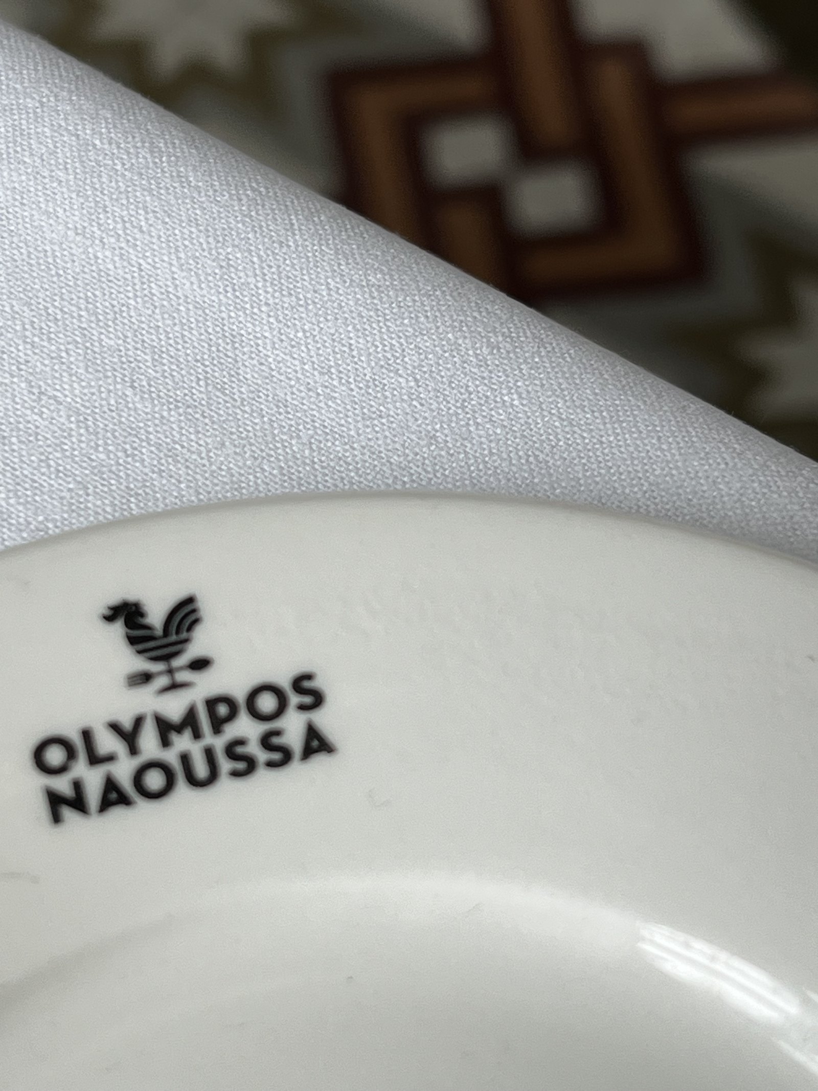 A white plate stamped OLYMPOS NAOUSSA with a rooster above the lettering, a folded napkin and a patterned cloth behind it