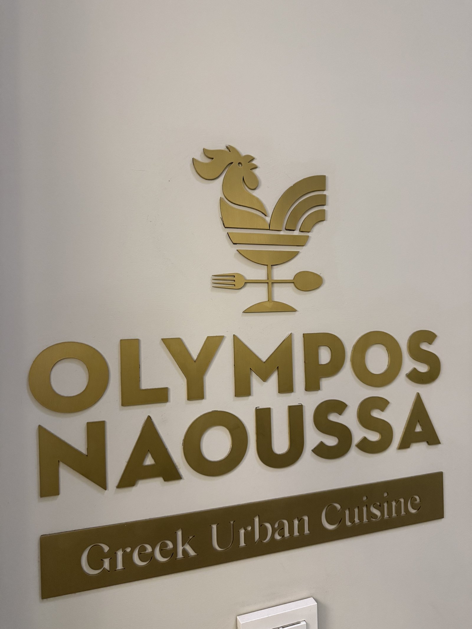 A gold sign reading OLYMPOS NAOUSSA, Greek Urban Cuisine, with a rooster above the lettering