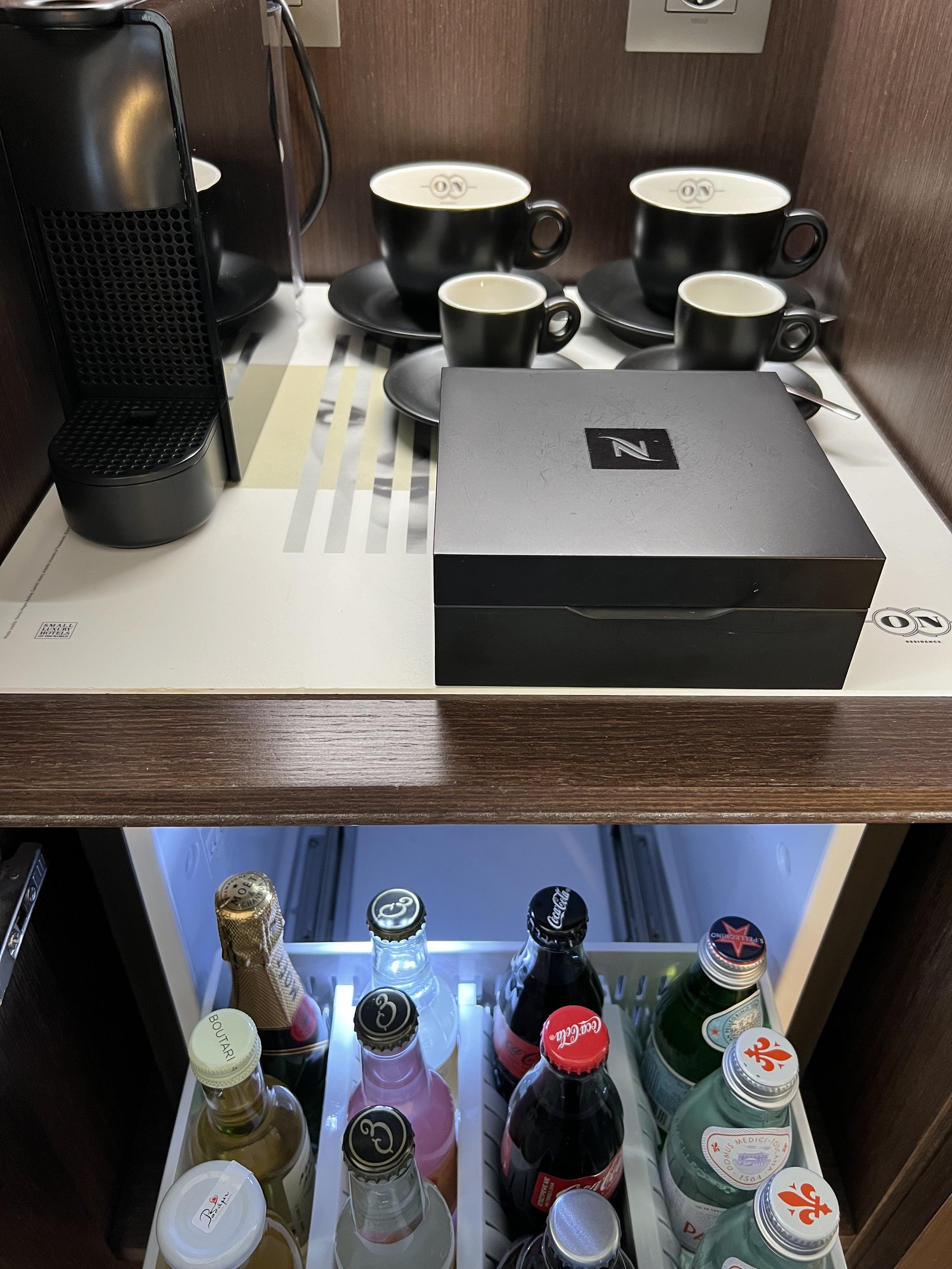 A Nespresso machine with cups and a black box above an open minibar drawer of Greek bottles