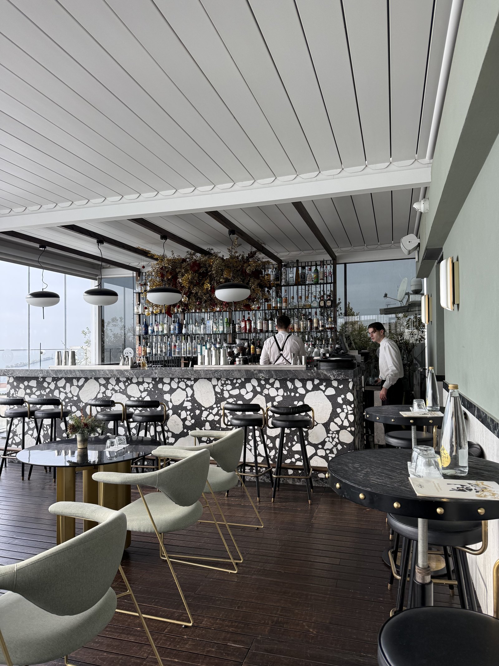 A rooftop bar counter with pale green stools, staff behind it and the sea through the windows