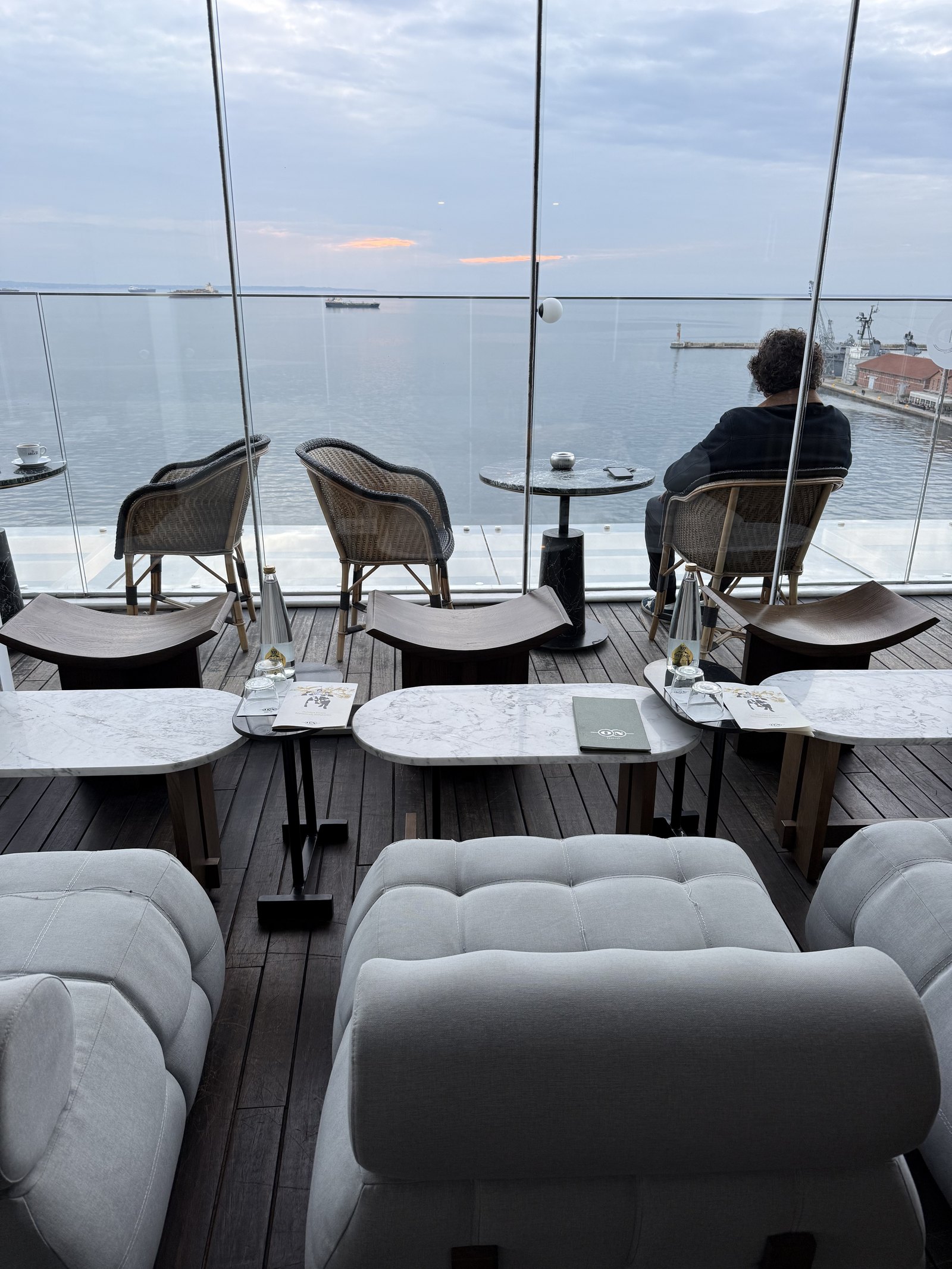 Cane chairs and low tables on a rooftop terrace at dusk, the gulf and the far shore beyond the glass