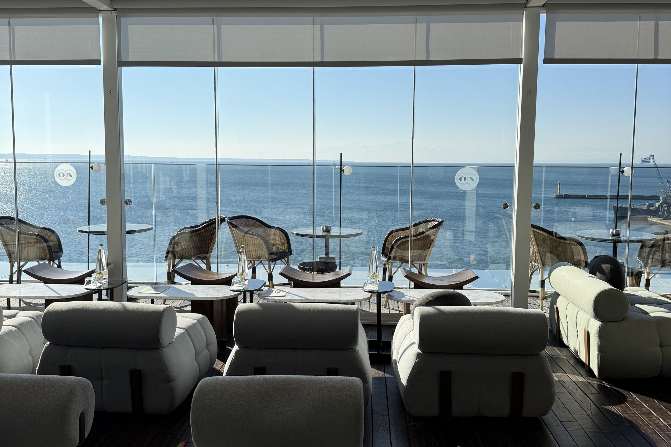 A rooftop bar behind a wall of glass, cane chairs and low cream seating facing the flat blue water of the Thermaic Gulf