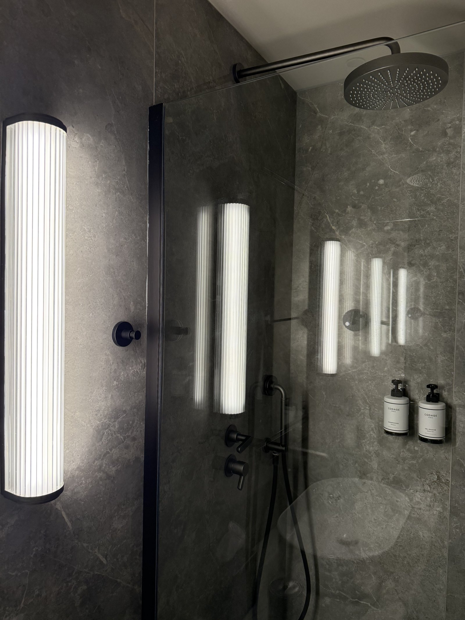A walk-in shower in grey marble with fluted vertical light tubes beside it