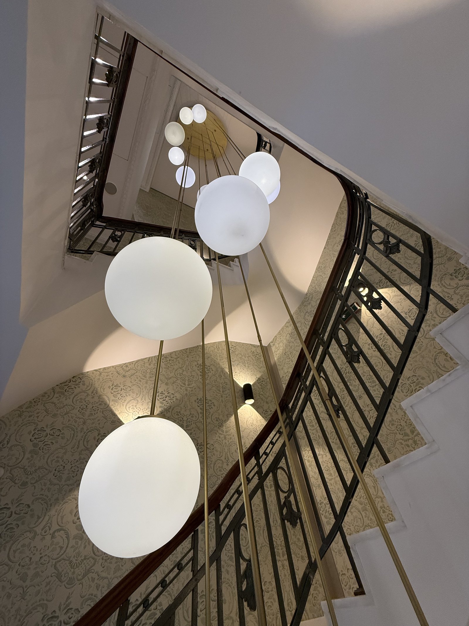 A stairwell seen from above, a cluster of white globe lights hanging down the well past an iron balustrade